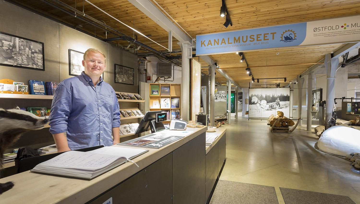 Bildet viser en mann som står bak resepsjonsdisken på Kanalmuseet. Han smiler mot kameraet. Rundt ham er brosjyrer, bøker og en datamaskin. I bakgrunnen kan man se museums utstillinger med bilder, båter og tømmerstokker.