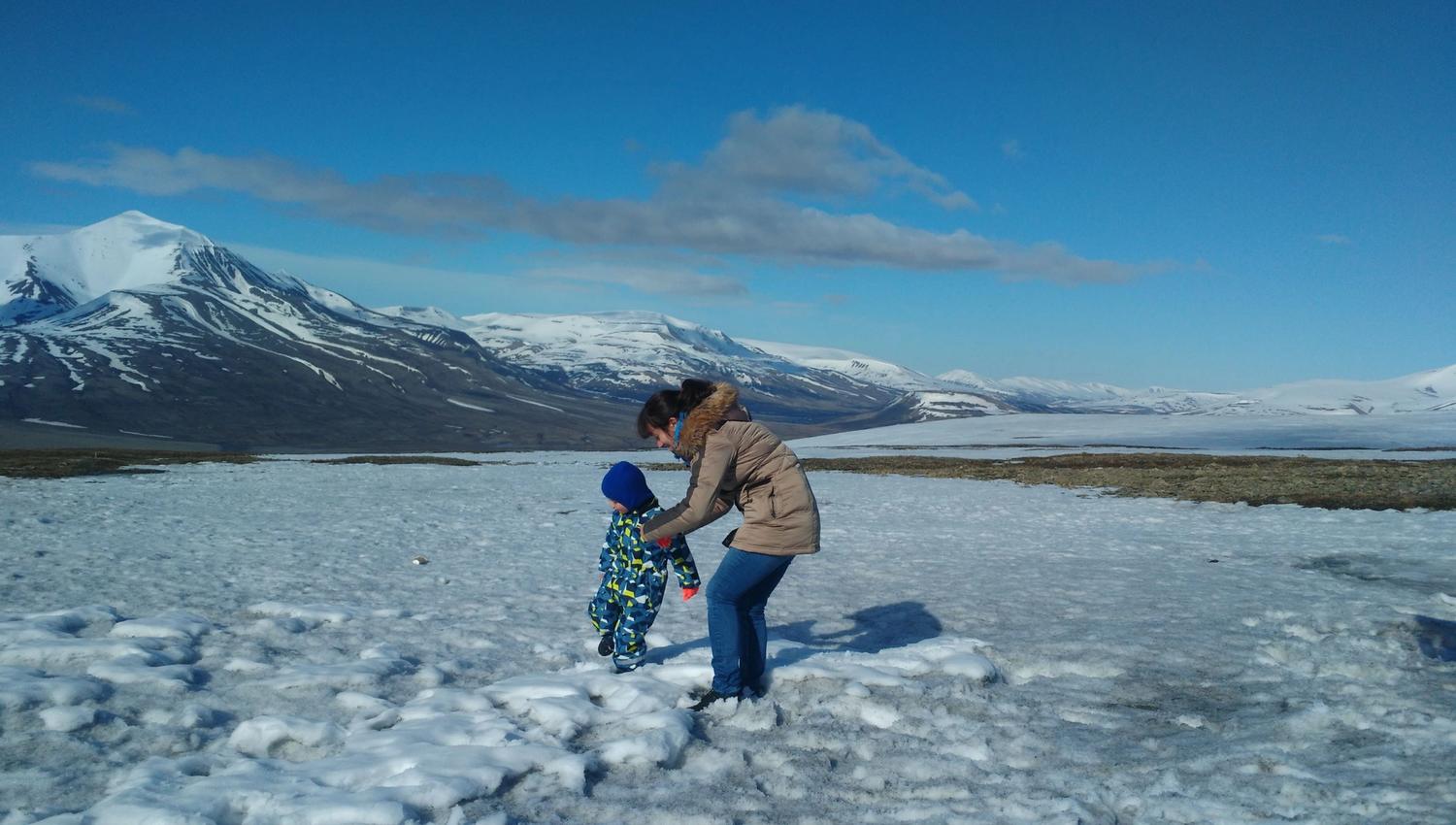A person holding a small person on their feet on the snow with snowy mountains in the background