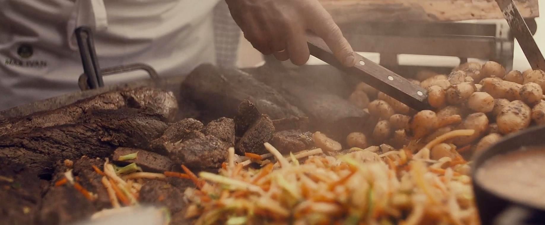 Nærbilde av grilling med kjøtt, poteter og grønnsaker på en stor grillplate. En person bruker grilltang til å vende maten.