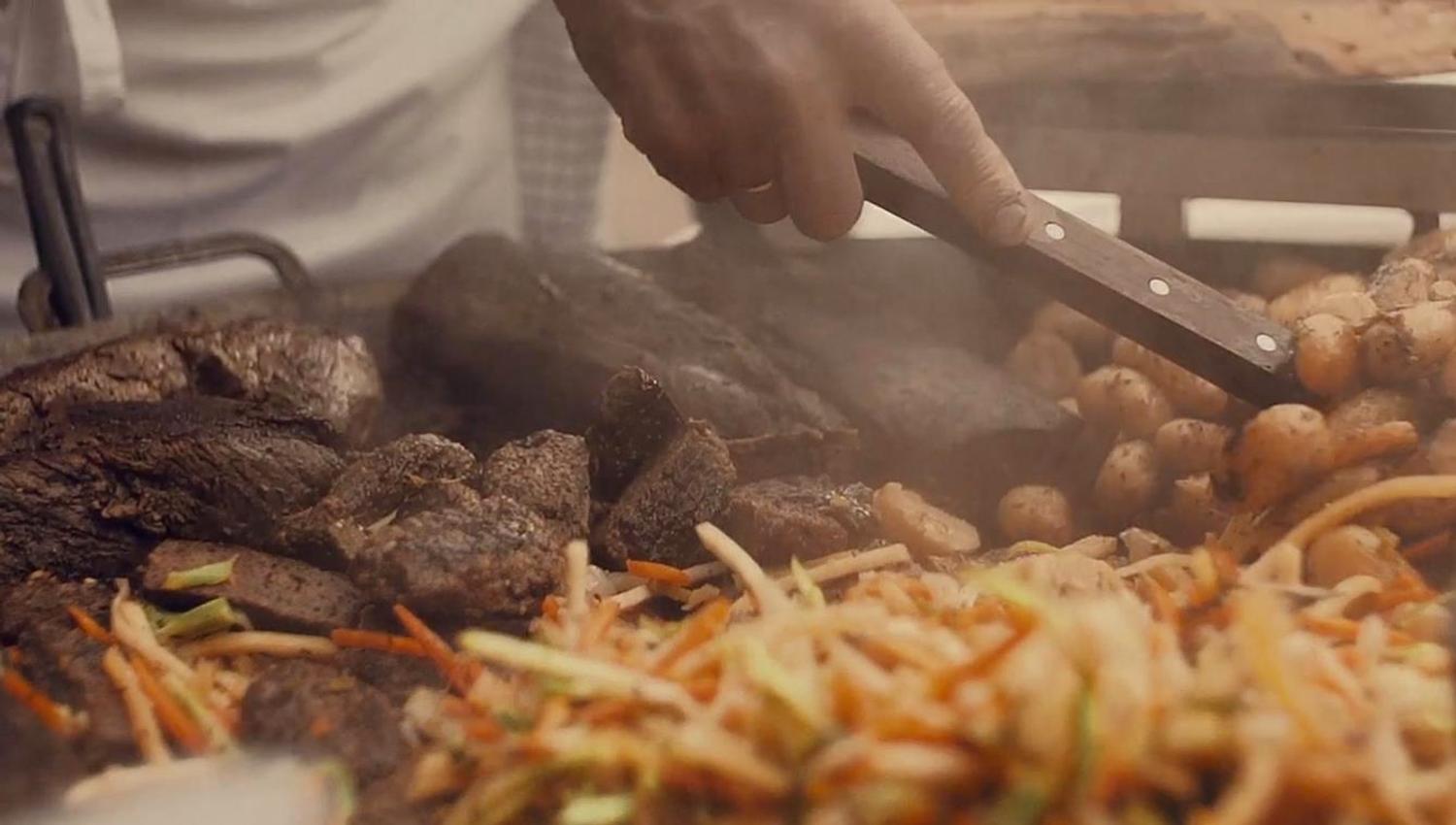 Nærbilde av grilling med kjøtt, poteter og grønnsaker på en stor grillplate. En person bruker grilltang til å vende maten.