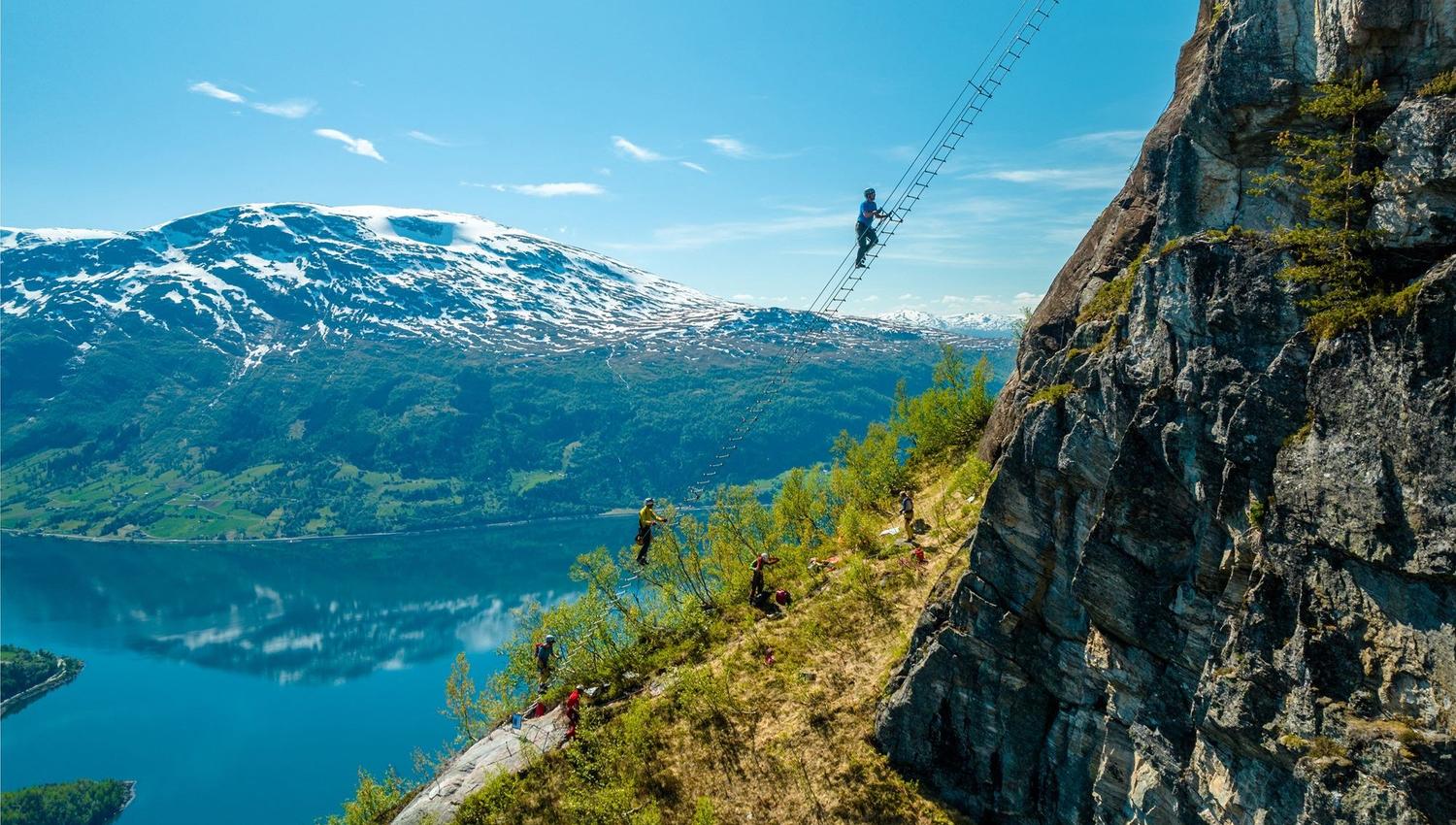 Ein person klatrar på ei luftig stige ved ein bratt fjellvegg, med fjord og snødekte fjell i bakgrunnen..