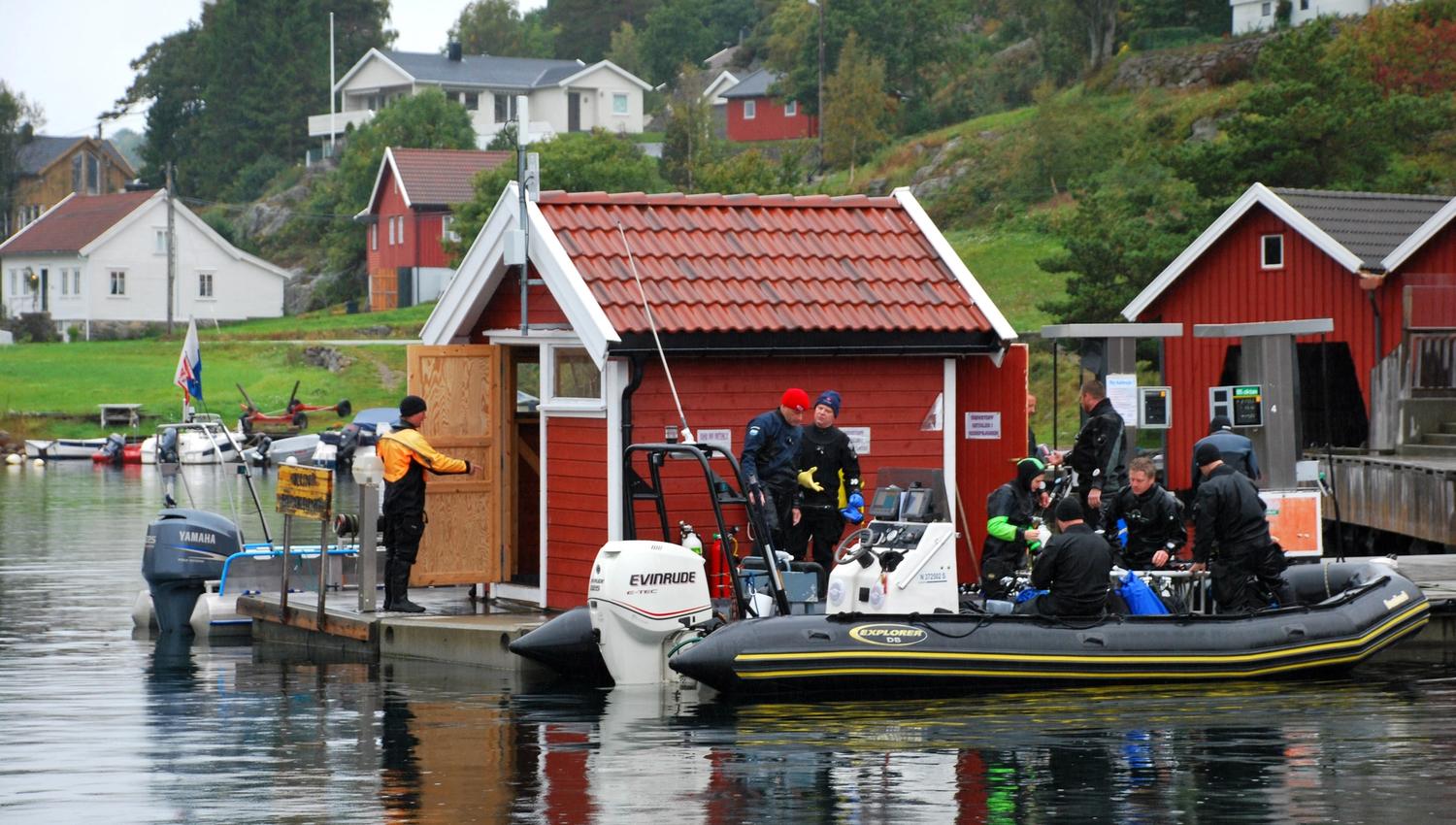 Picture of a red shed on a pier with at least 8 divers getting ready to board a black boat at Tregde