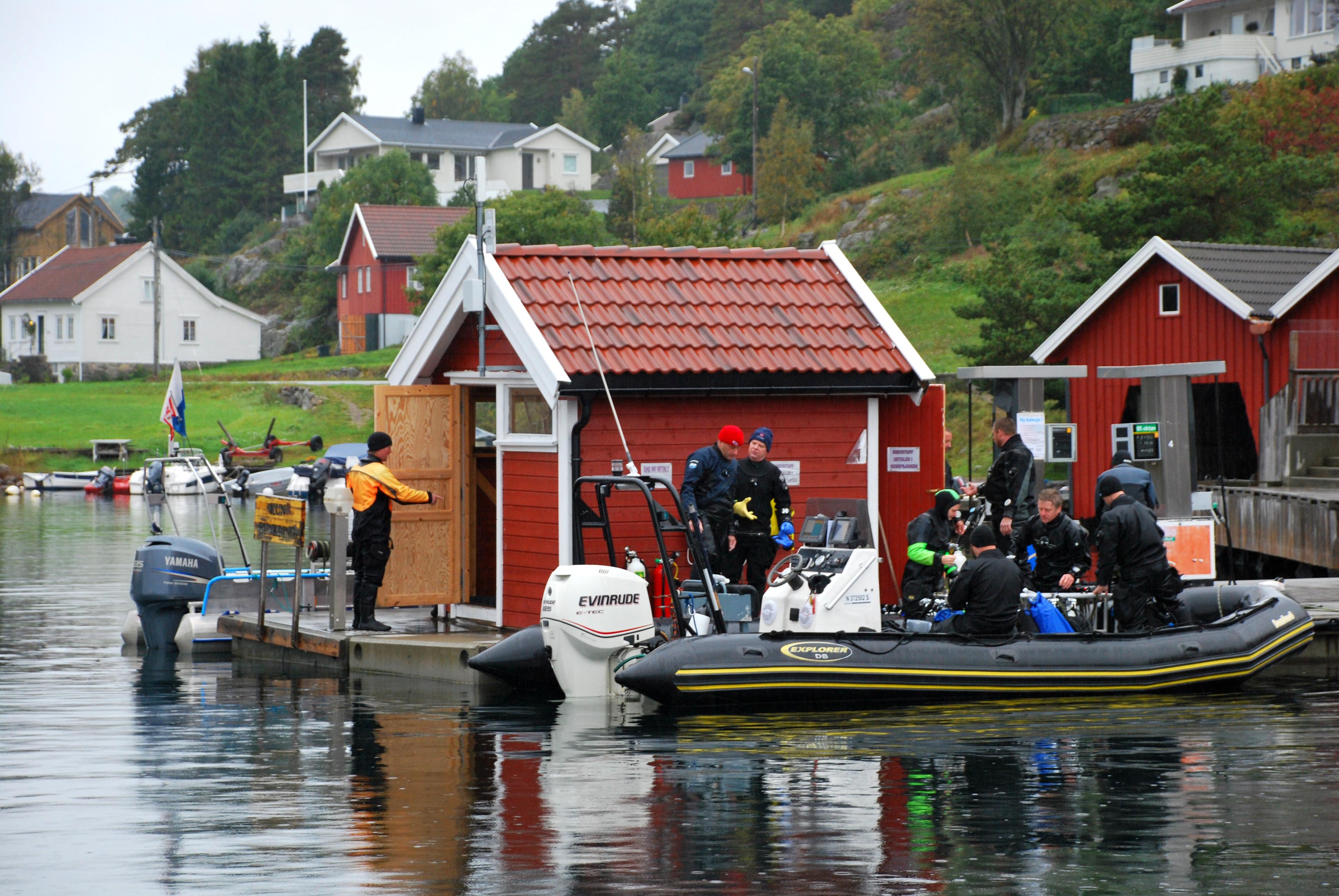 Picture of a red shed on a pier with at least 8 divers getting ready to board a black boat at Tregde