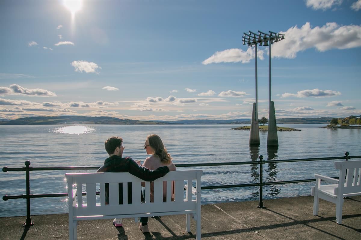 To personer sitter på en benk ved vannet, med utsikt mot skulpturer og en lys himmel.
