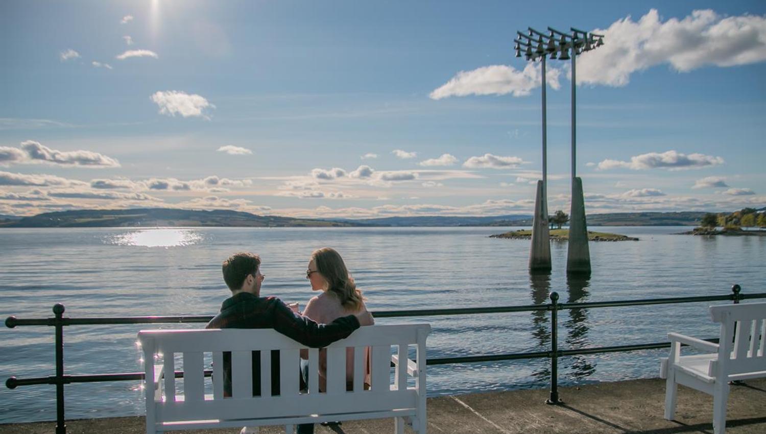 To personer sitter på en benk ved vannet, med utsikt mot skulpturer og en lys himmel.