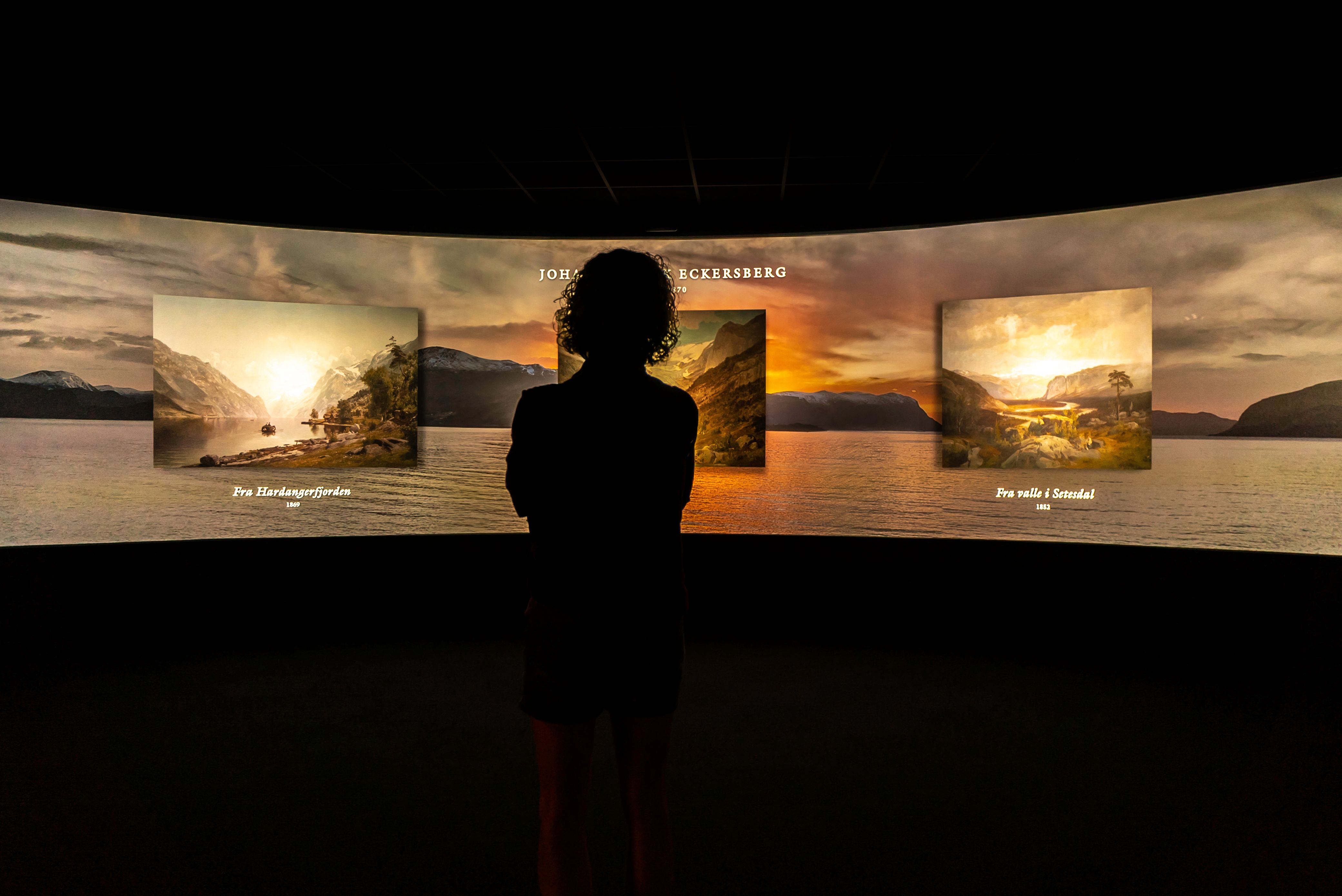 A woman gazing at the large panorama screen displaying various pieces of artwork by norwegian painters.