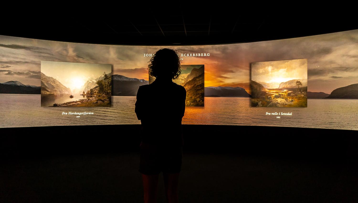 A woman gazing at the large panorama screen displaying various pieces of artwork by norwegian painters.