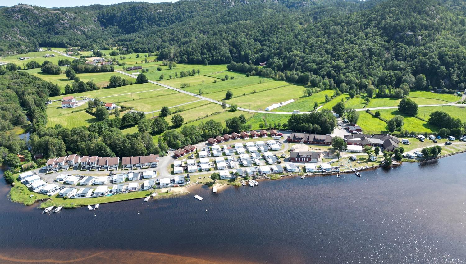 Aerial view of Solstrand Camping. Many caravans and cabins are situated along the waterfront. Behind the campsite are large fields, with forest furth