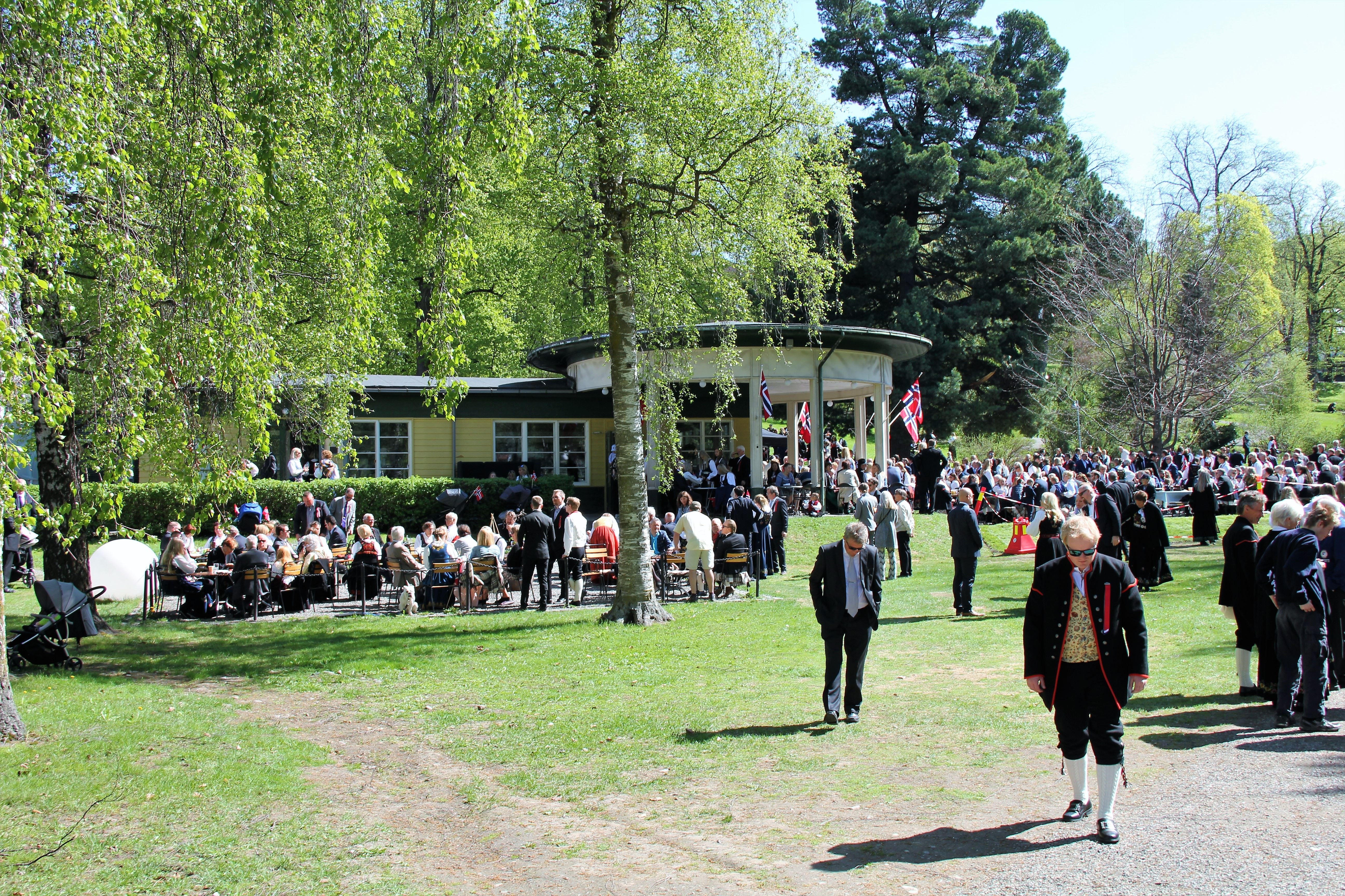 Stor folkemengde i en grønn park rundt en paviljong med norske flagg på en solrik dag, mange i finstøy..