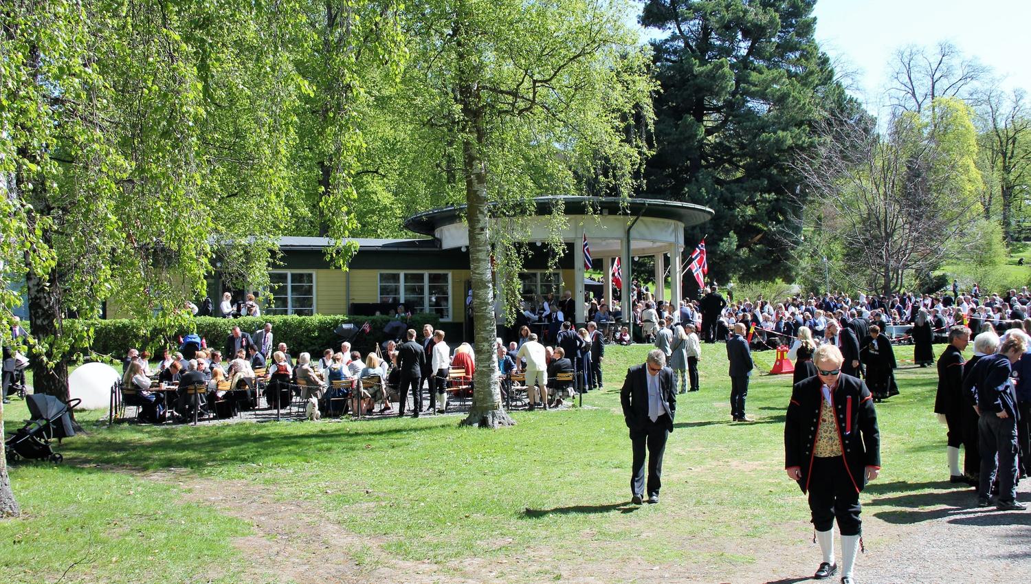 Stor folkemengde i en grønn park rundt en paviljong med norske flagg på en solrik dag, mange i finstøy..