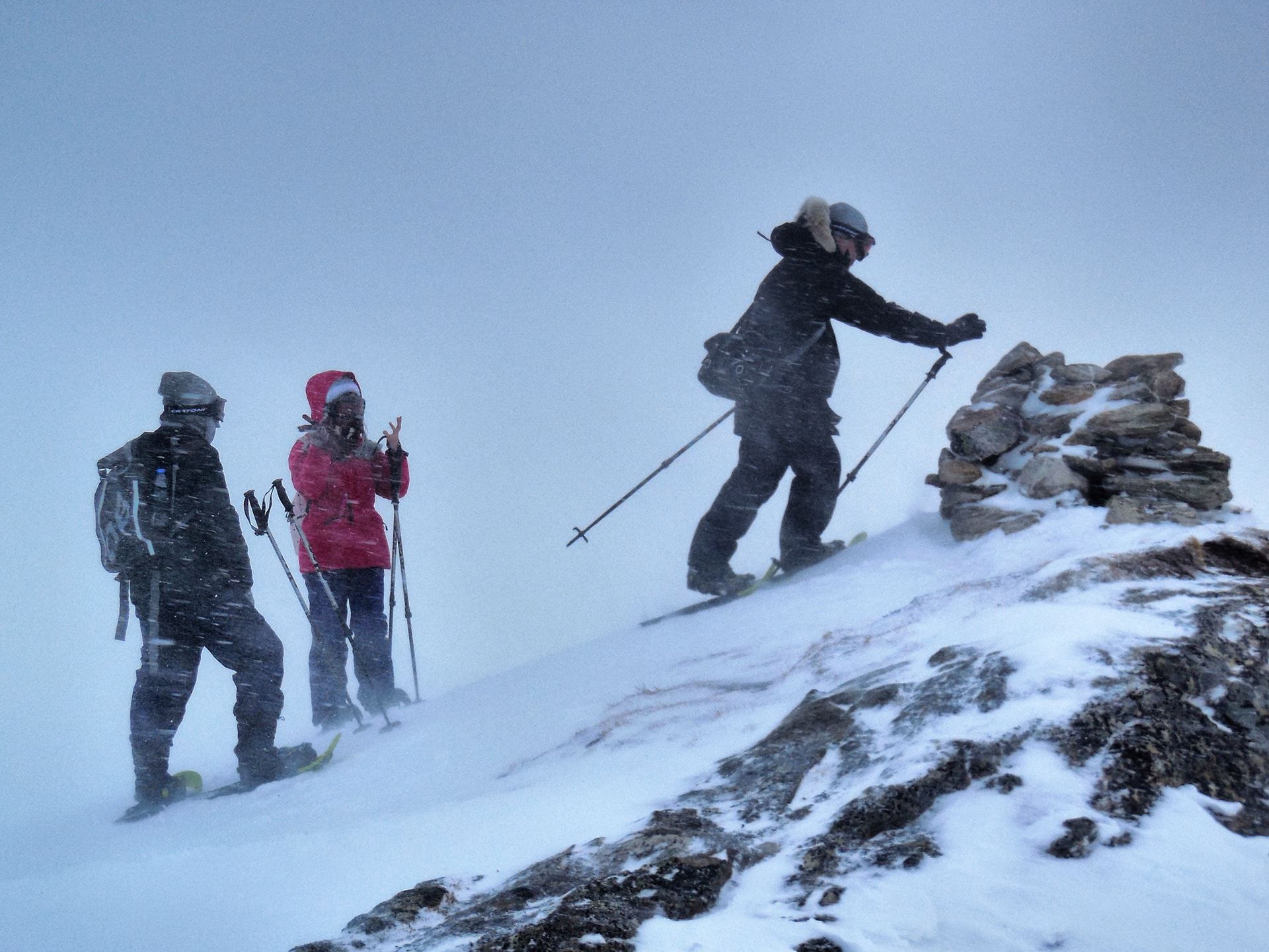 Three people in winter gear ascending a snowy mountain in windy conditions, one placing a stone on a cairn.