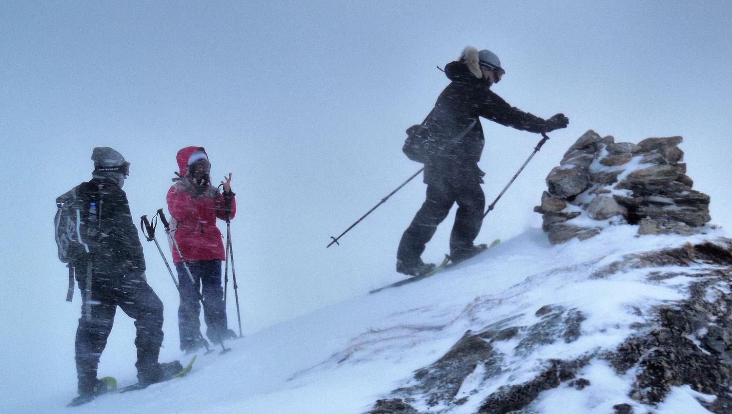 Three people in winter gear ascending a snowy mountain in windy conditions, one placing a stone on a cairn.
