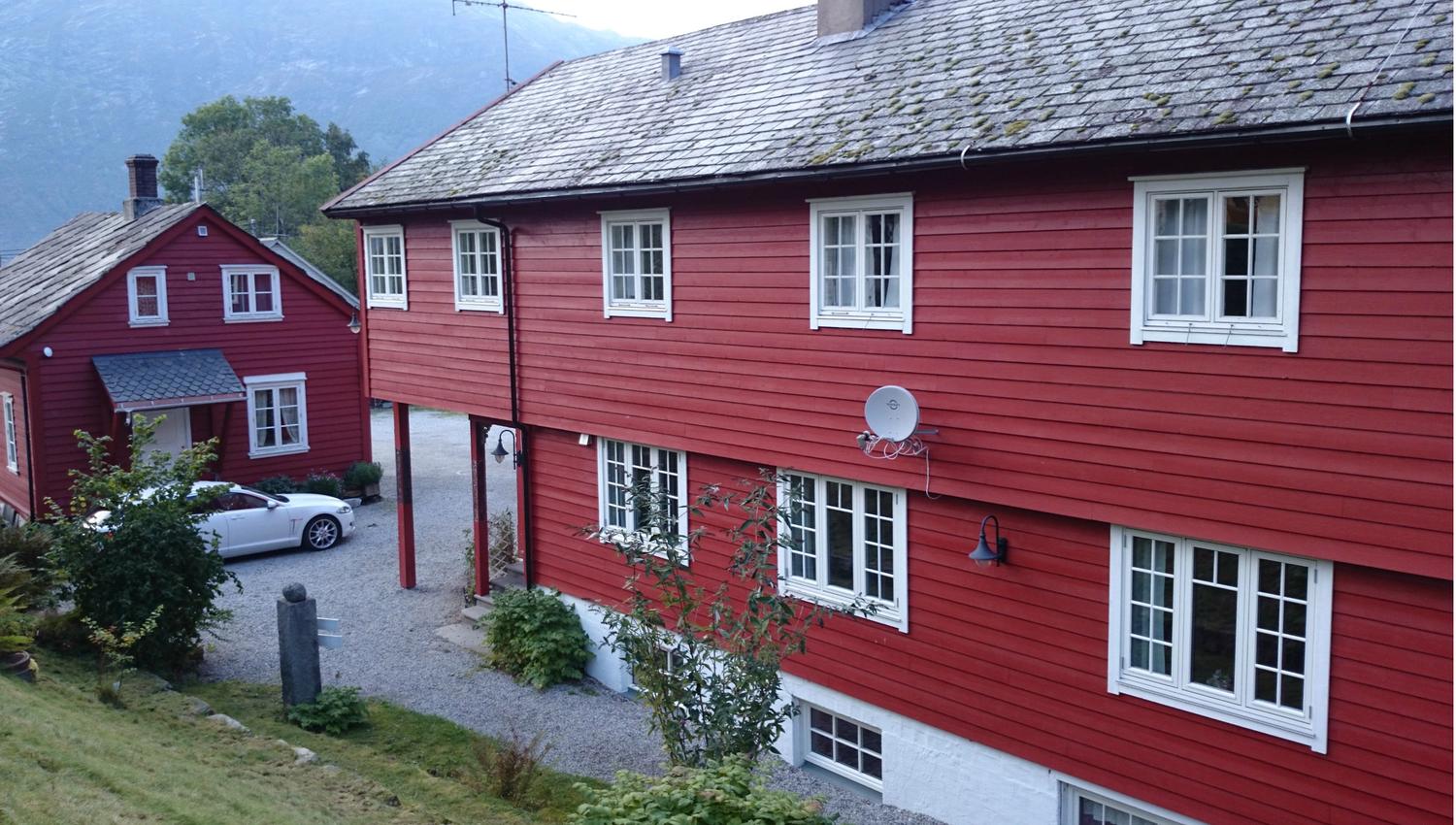 Raudmåla hovudbygg og tun på Ullensvang Gjesteheim, omkransa av fjell, frukthagar og tradisjonell vestlandsidyll.