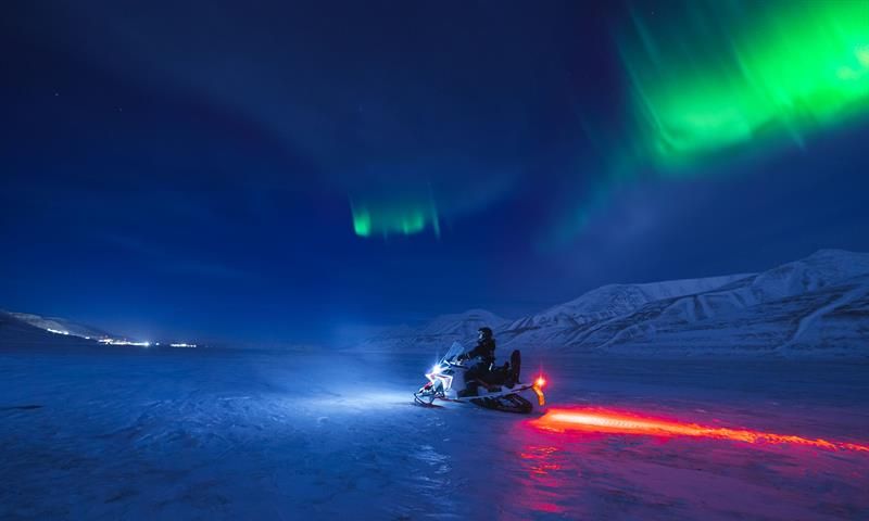 Nordlys over snødekte fjell med en snøscooter kjørende forbi