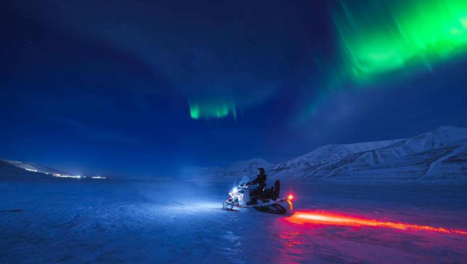 Nordlys over snødekte fjell med en snøscooter kjørende forbi