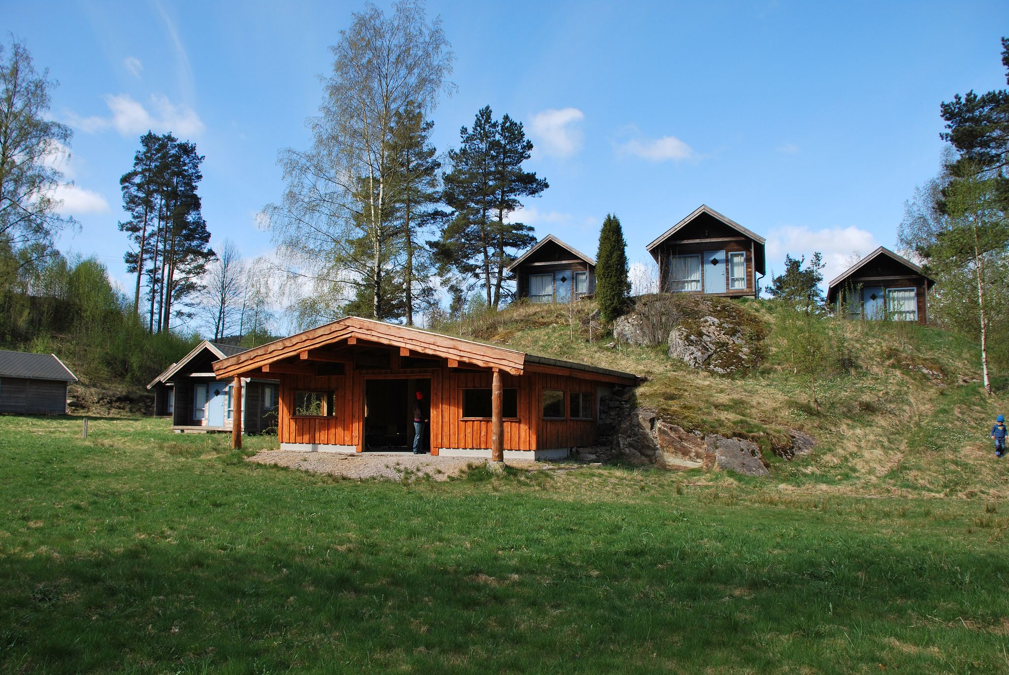 Oransj hytte midt på bildet, med flere små brune hytter bak på en haug. Få trær, og blåhimmel med noen få skyer. Gresset foran hyttene er grønt