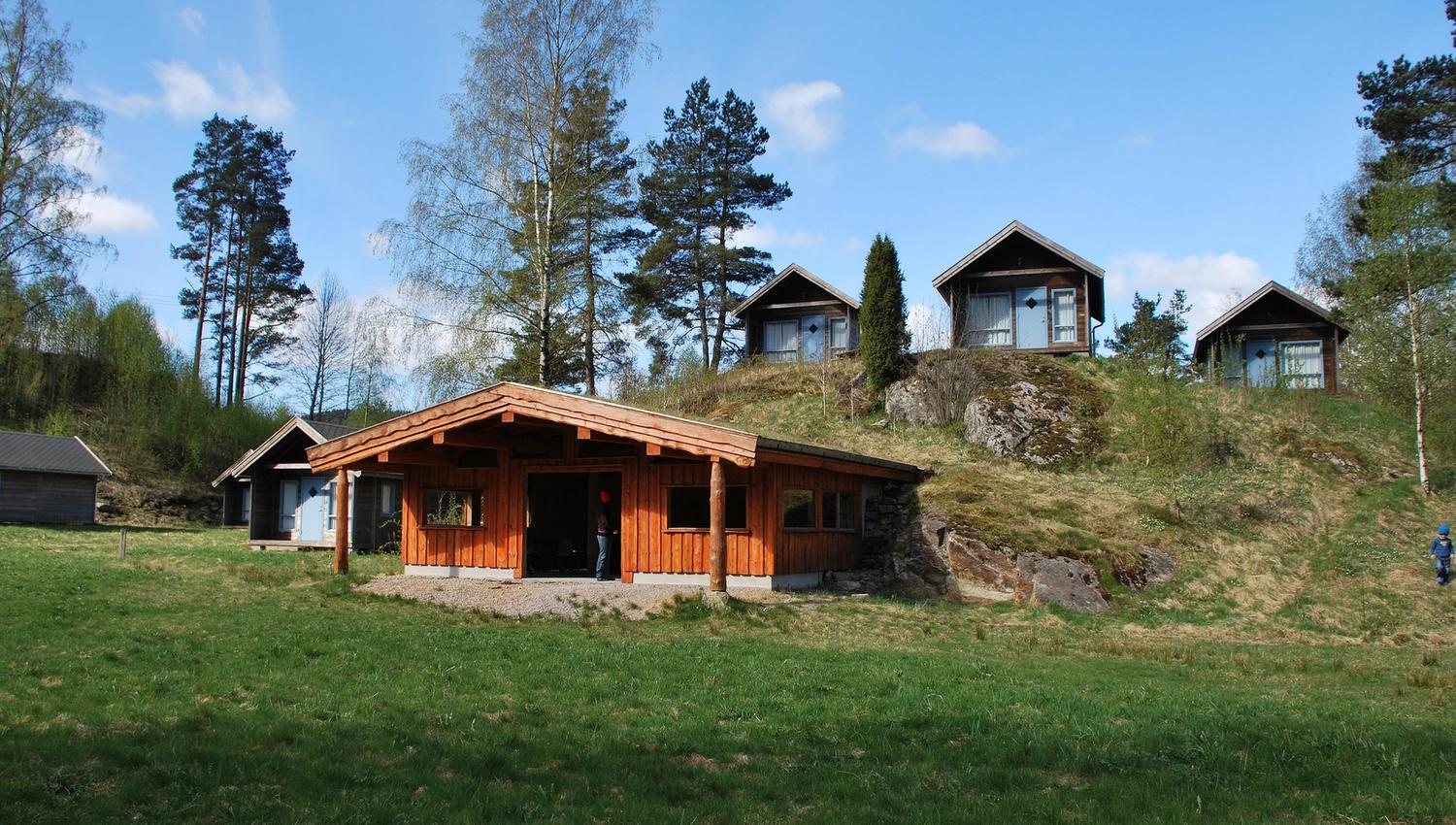Oransj hytte midt på bildet, med flere små brune hytter bak på en haug. Få trær, og blåhimmel med noen få skyer. Gresset foran hyttene er grønt