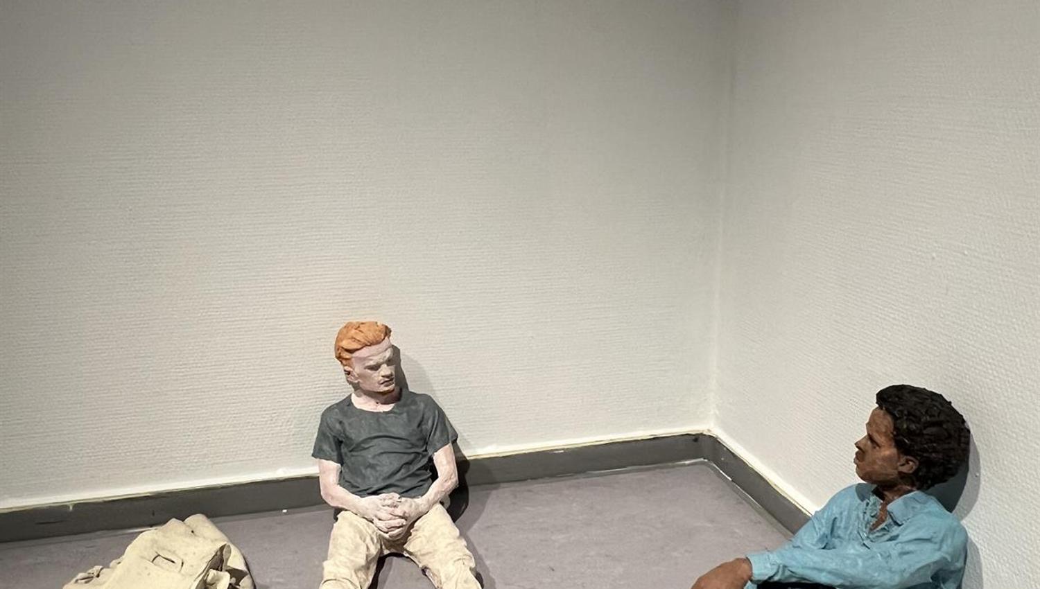 To skulpturfigurer sittende på gulvet ved vegg med gjenstander rundt.