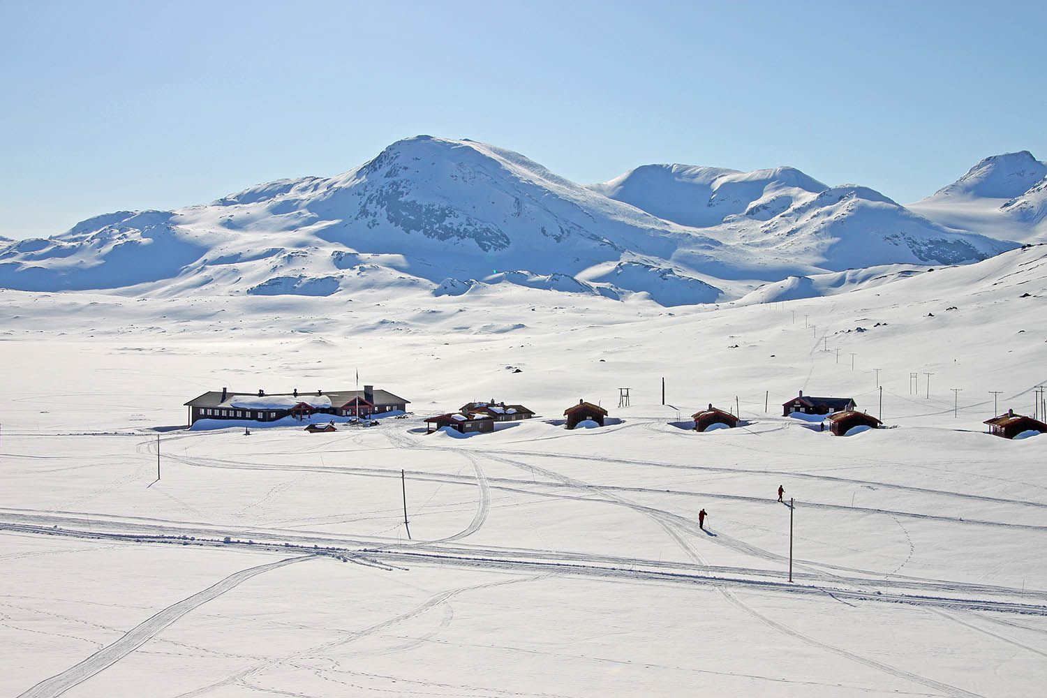 En liten fjellstue og et knippe hytter i et nedsnødd høyfjellslandskap fra fugleperspektivet. Skispor og skigåere utenfor hytta.