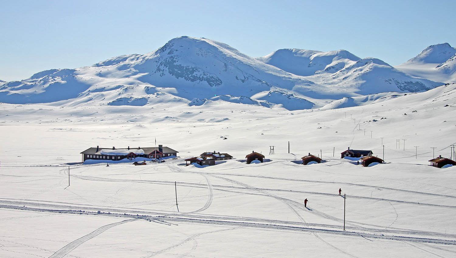 En liten fjellstue og et knippe hytter i et nedsnødd høyfjellslandskap fra fugleperspektivet. Skispor og skigåere utenfor hytta.