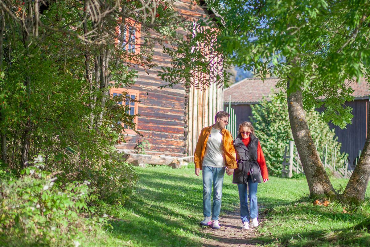 Tre personer går på en gressplen ved siden av en trebygning omgitt av grønne trær.