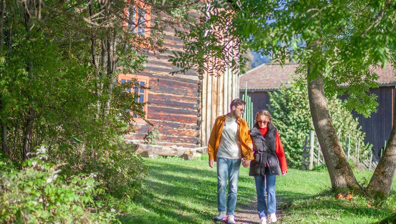 Tre personer går på en gressplen ved siden av en trebygning omgitt av grønne trær.