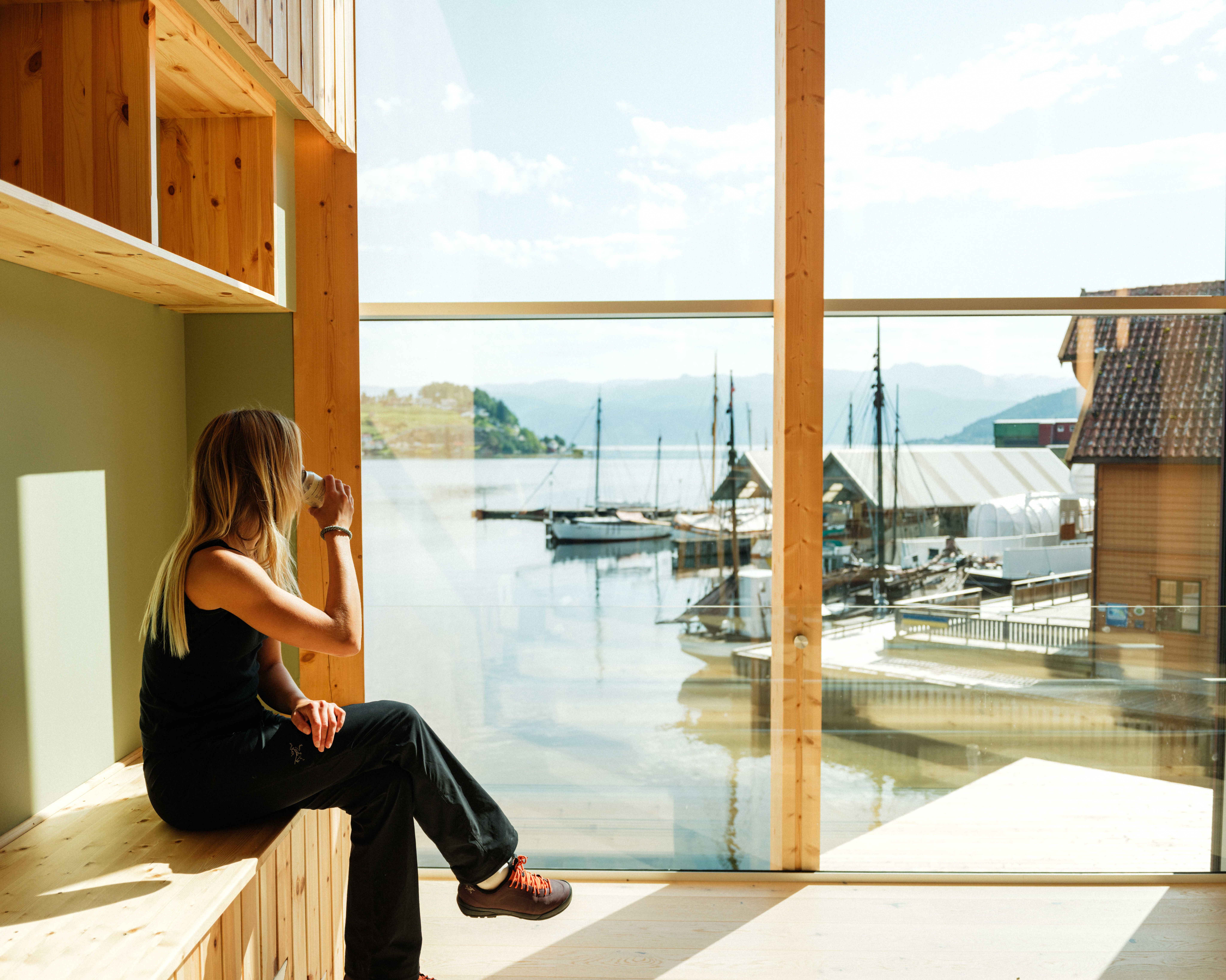 Woman enjoying a coffee break with a stunning view of the fjord and traditional boats at the Hardanger Maritime Centre.