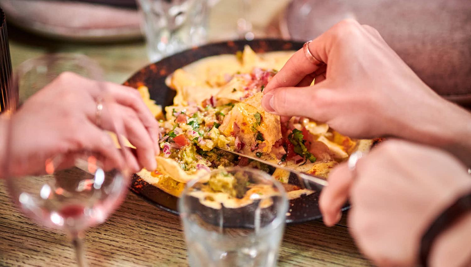 Personer deler en tallerken nachos ved et bord med vin og vann.