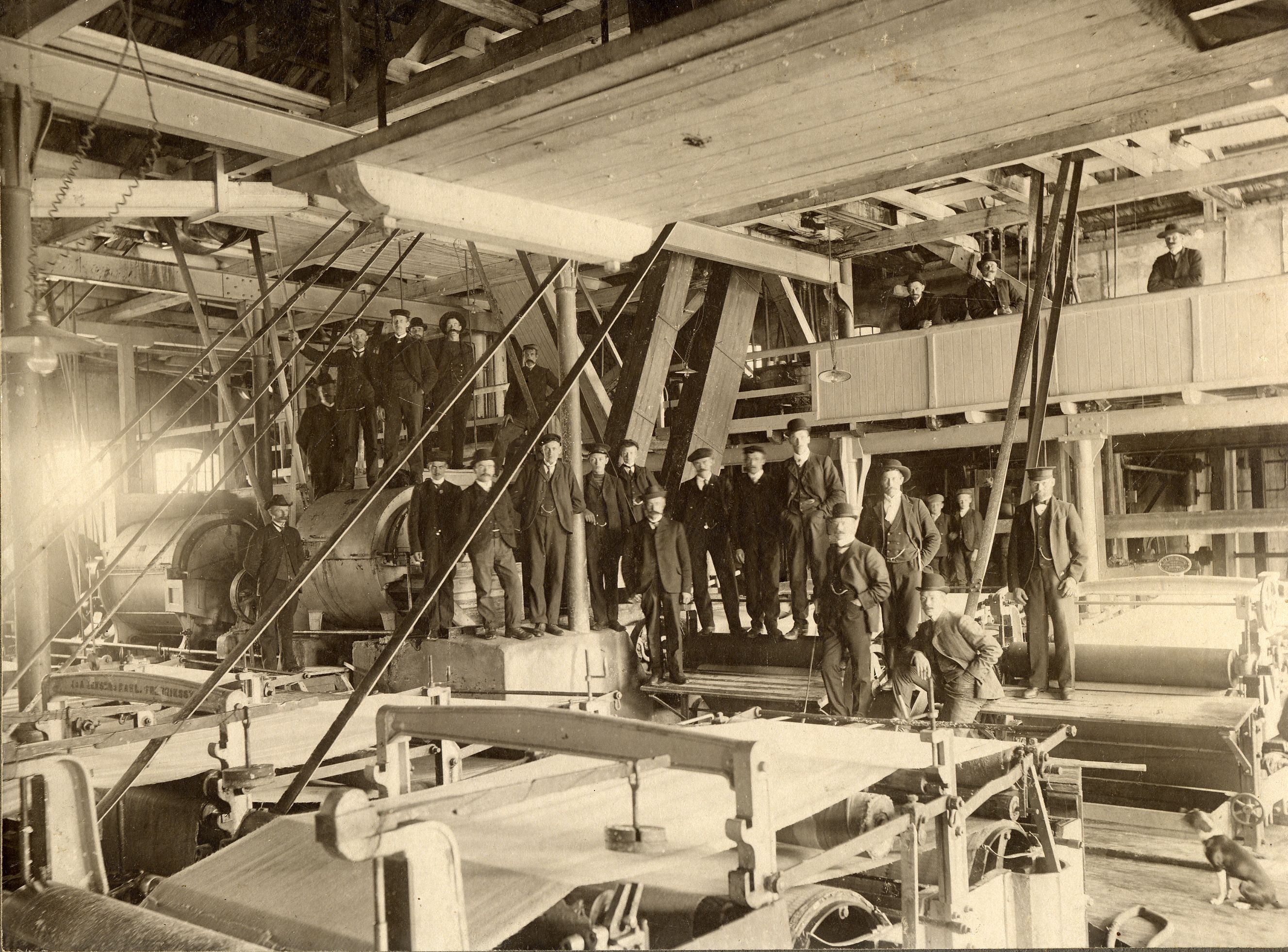 Bildet er et gammelt svart-hvitt foto som viser en stor gruppe menn inne i en fabrikk eller et sagbruk. De står blant store maskiner og bjelker, og mange har på seg hatter og arbeidsklær. Lyset kommer inn fra venstre side.