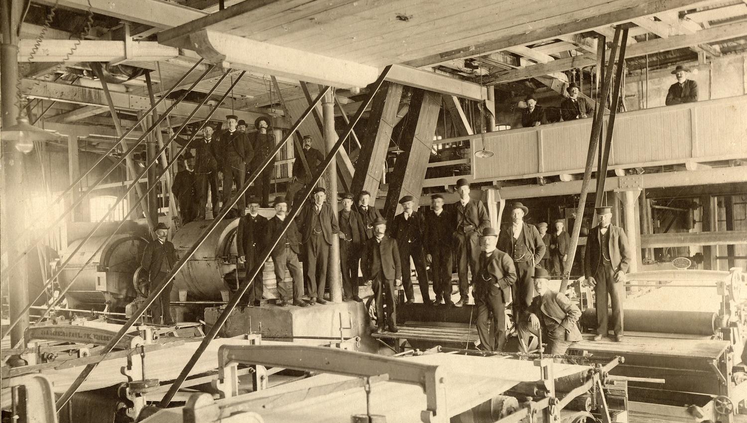 Bildet er et gammelt svart-hvitt foto som viser en stor gruppe menn inne i en fabrikk eller et sagbruk. De står blant store maskiner og bjelker, og mange har på seg hatter og arbeidsklær. Lyset kommer inn fra venstre side.