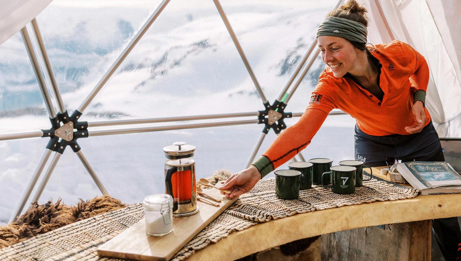 A smiling woman in orange and blue attire preparing tea on a triangular table inside a geodesic dome with mountain views.