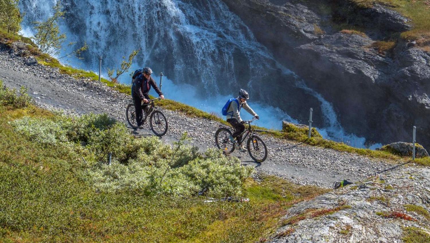 To syklistar på Rallarvegen passerer ein foss i Hardangerfjorden-området.