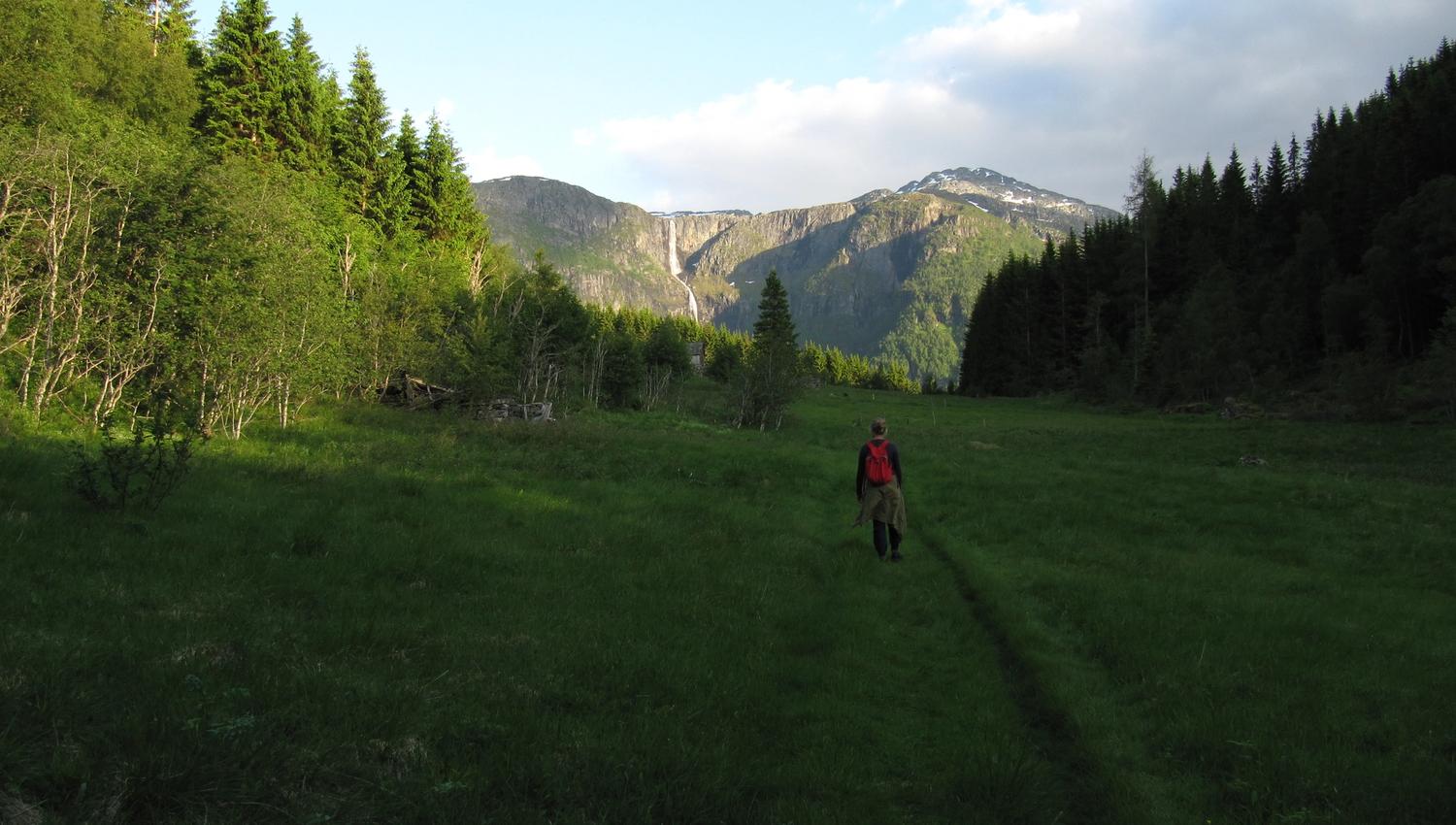 Vandrar med raud sekk på stien gjennom dalen på Hovden rundt med fjell og fossar i horisonten.