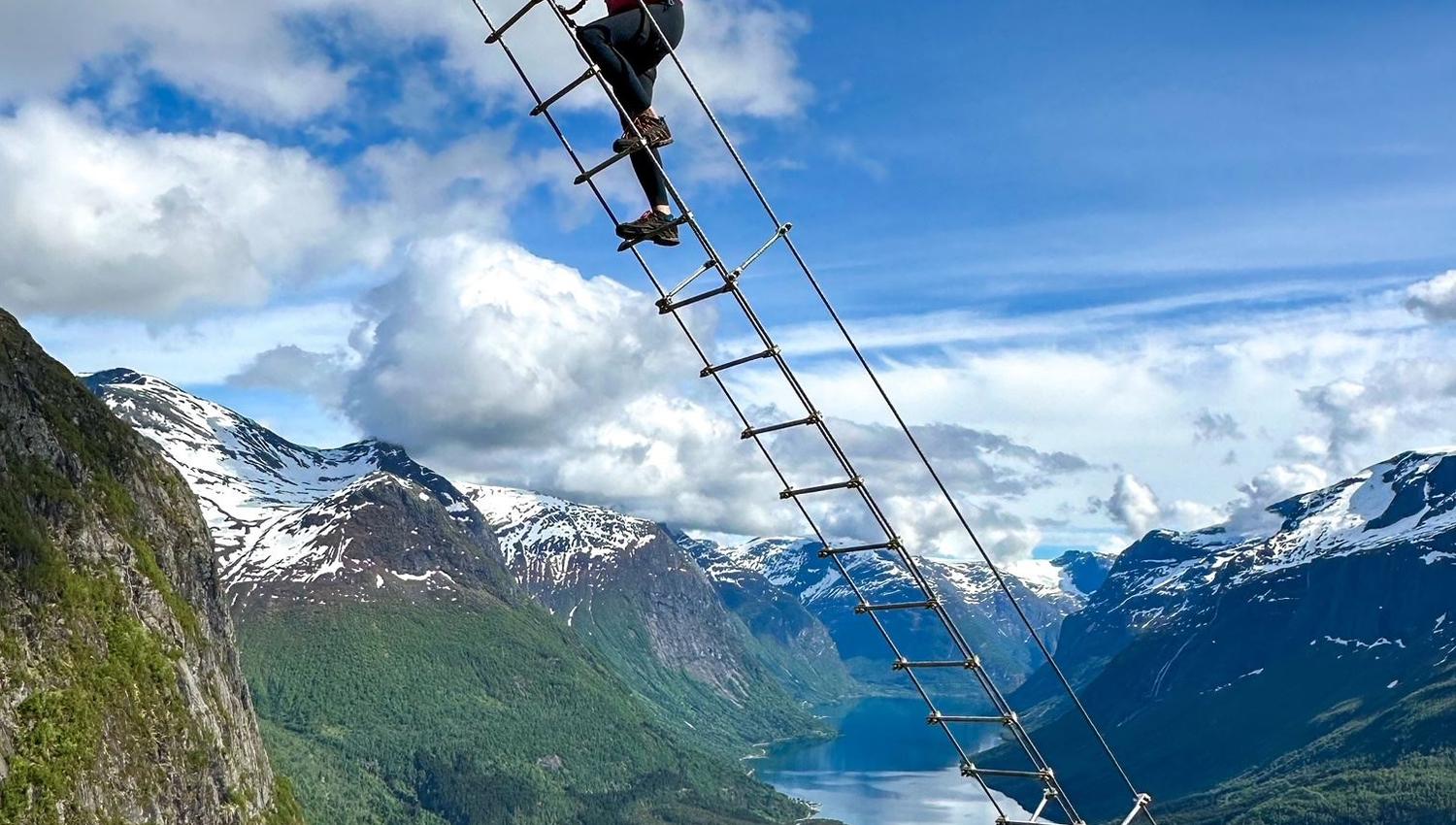 Ein person klatrar ei taustige høgt over ein grøn dal med fjord, snødekte fjell og blå himmel med skyer..