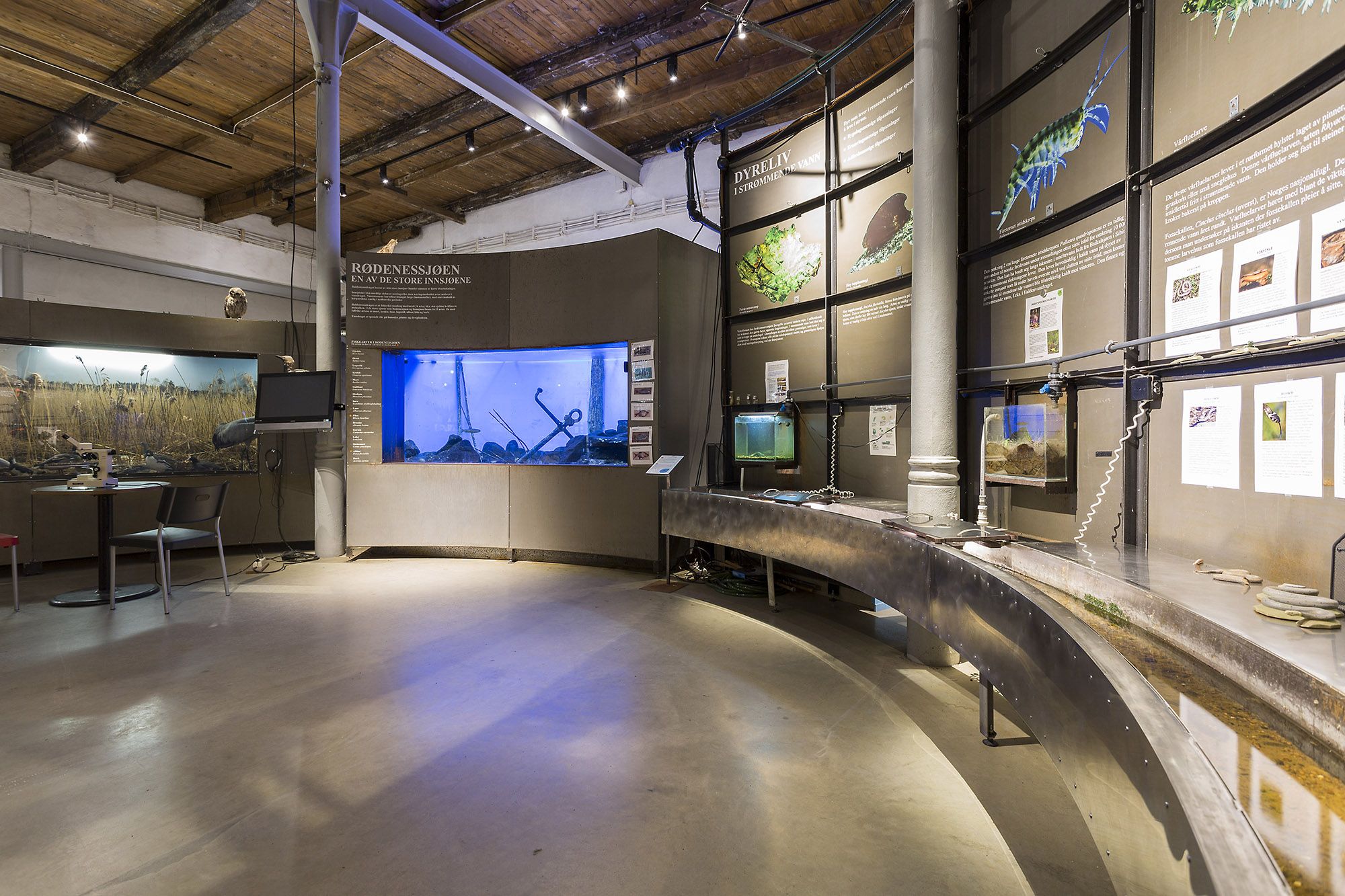 Bildet viser en utstilling inne på et museum. Langs veggene er det informasjonstavler, bilder av fisk og natur, og et stort akvarium med blått lys. Midt i rommet går en lang metall renne, trolig en del av en vann- eller kanalmodell.