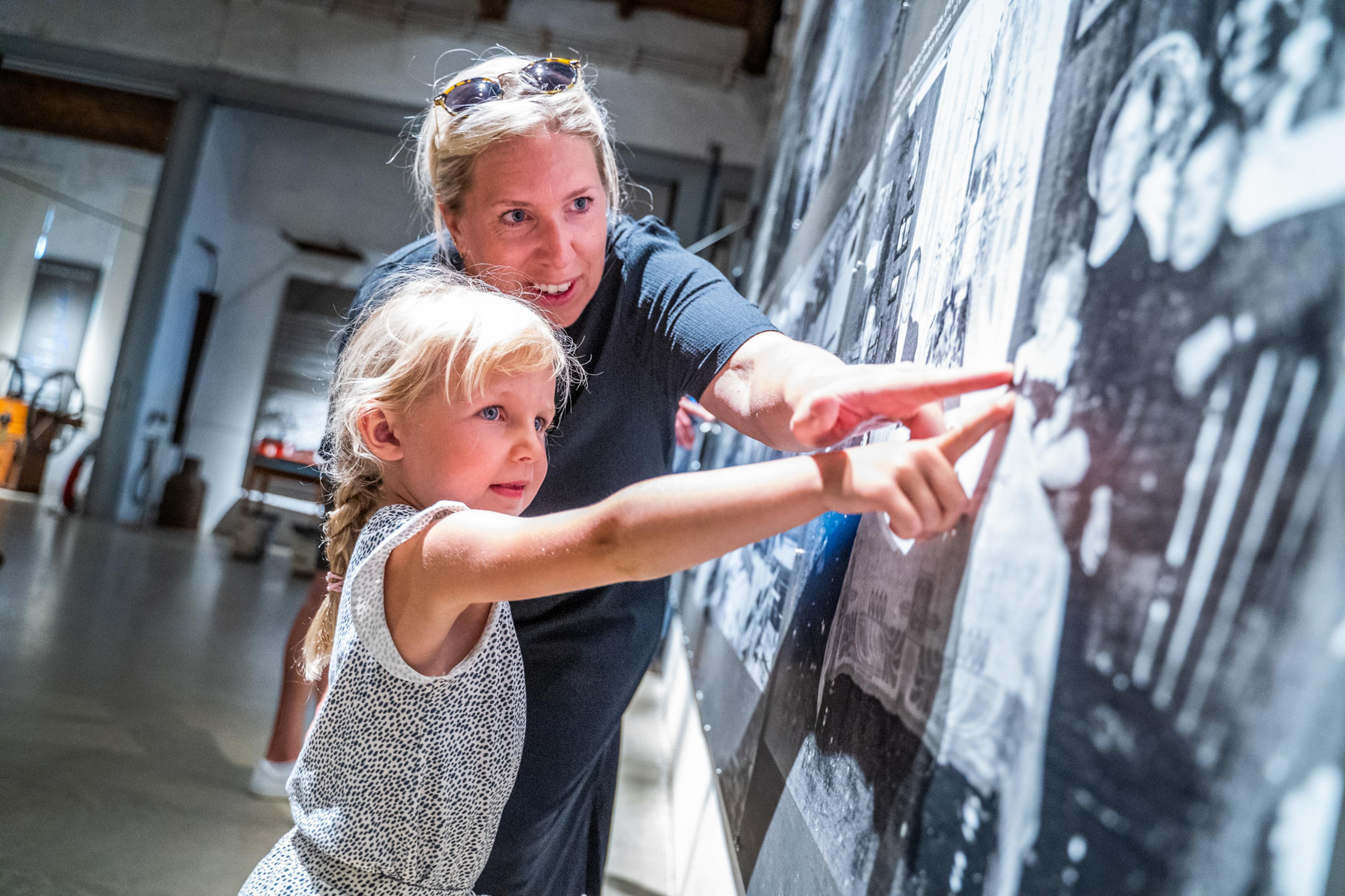 The picture shows a woman and a young girl looking at a large picture or exhibit on a wall. They are both pointing and smiling, and it looks like the 