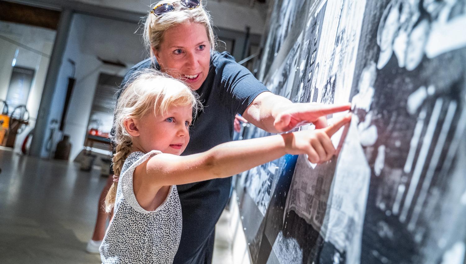 Bildet viser en kvinne og en liten jente som ser på et stort bilde eller en utstilling på en vegg. Begge peker og smiler, og det ser ut som kvinnen forklarer noe til jenta. De er inne i et lyst rom, de er på et museum.