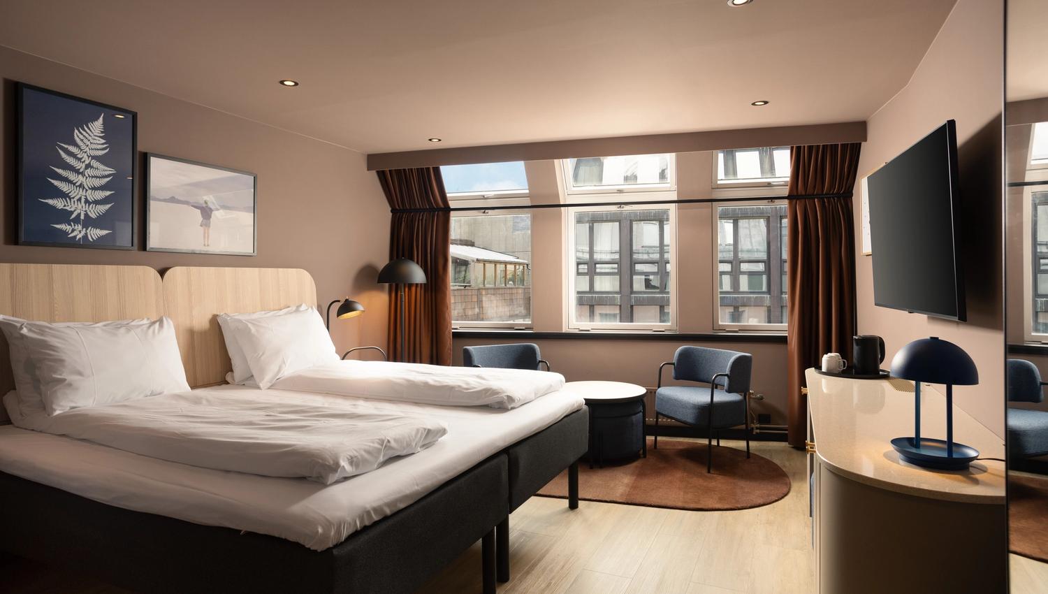 Hotel room with large double bed, TV on the wall, blue armchairs and brown curtains