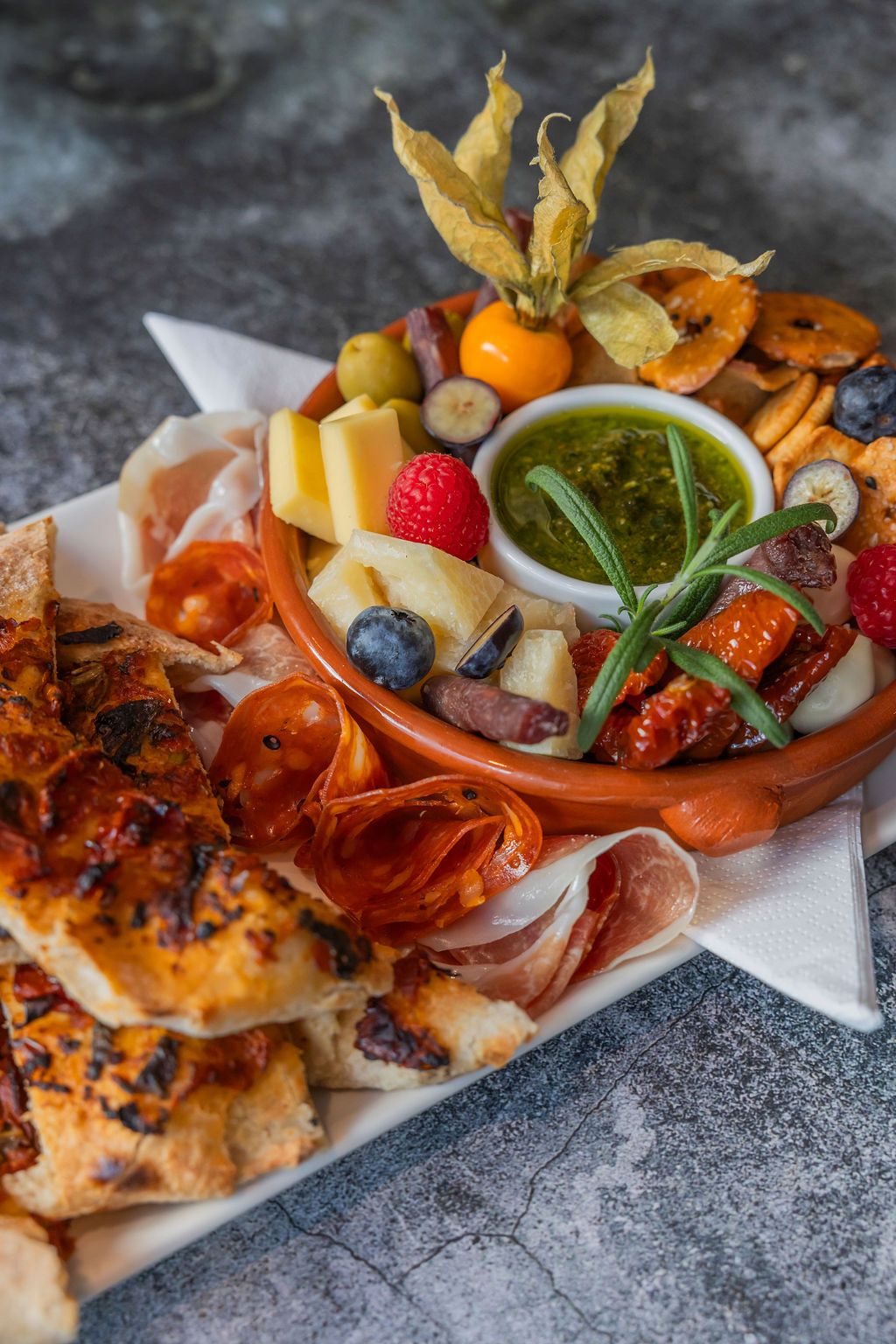 Matbilde tapastallerken, små pizzastykker, peperoni og skinke rullet sammen, liten skål med diverse snacks oppi. Blåbær, bringebær, physalis , ost, og en grønn dipp i midten.