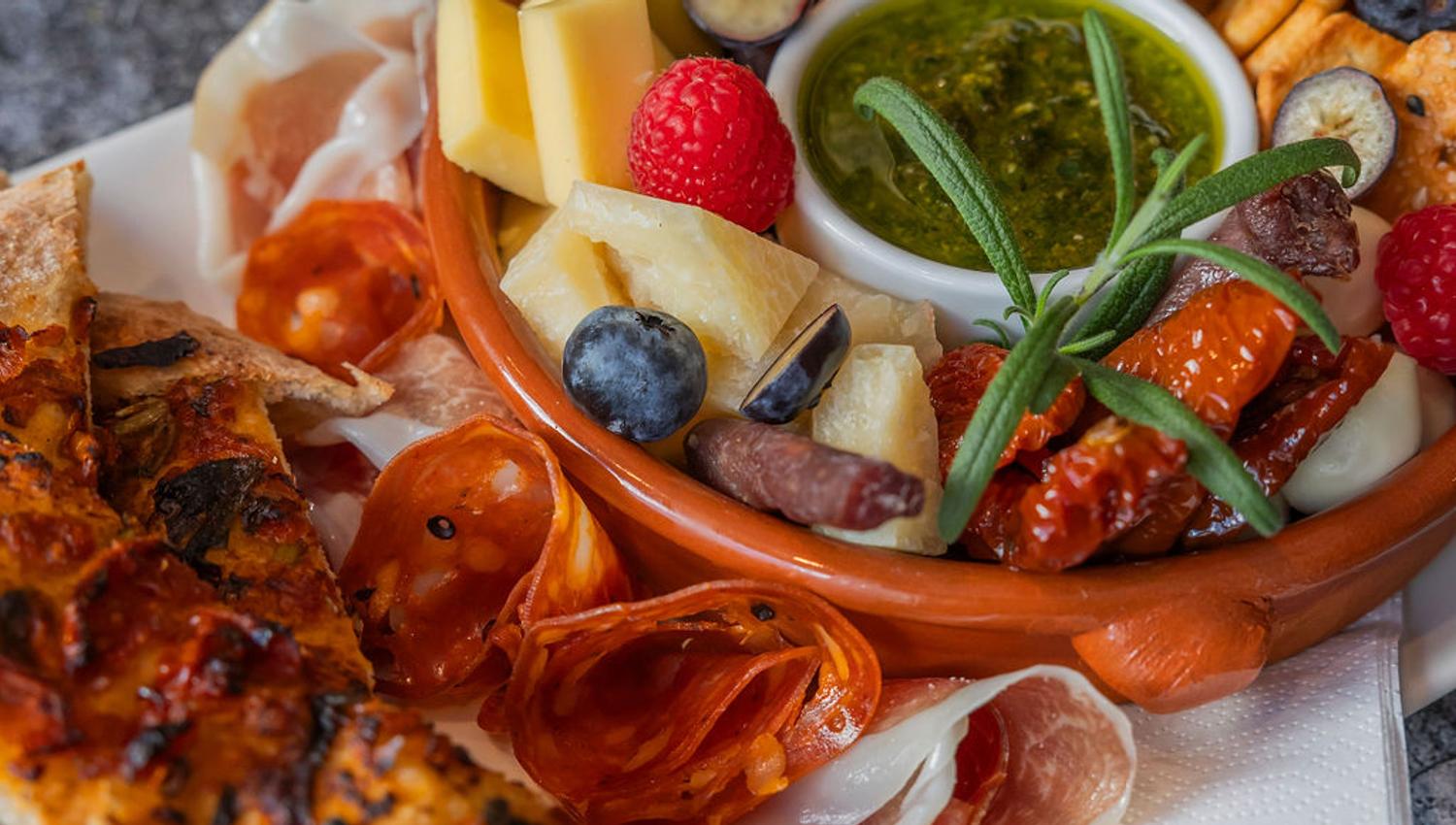 Matbilde tapastallerken, små pizzastykker, peperoni og skinke rullet sammen, liten skål med diverse snacks oppi. Blåbær, bringebær, physalis , ost, og en grønn dipp i midten.