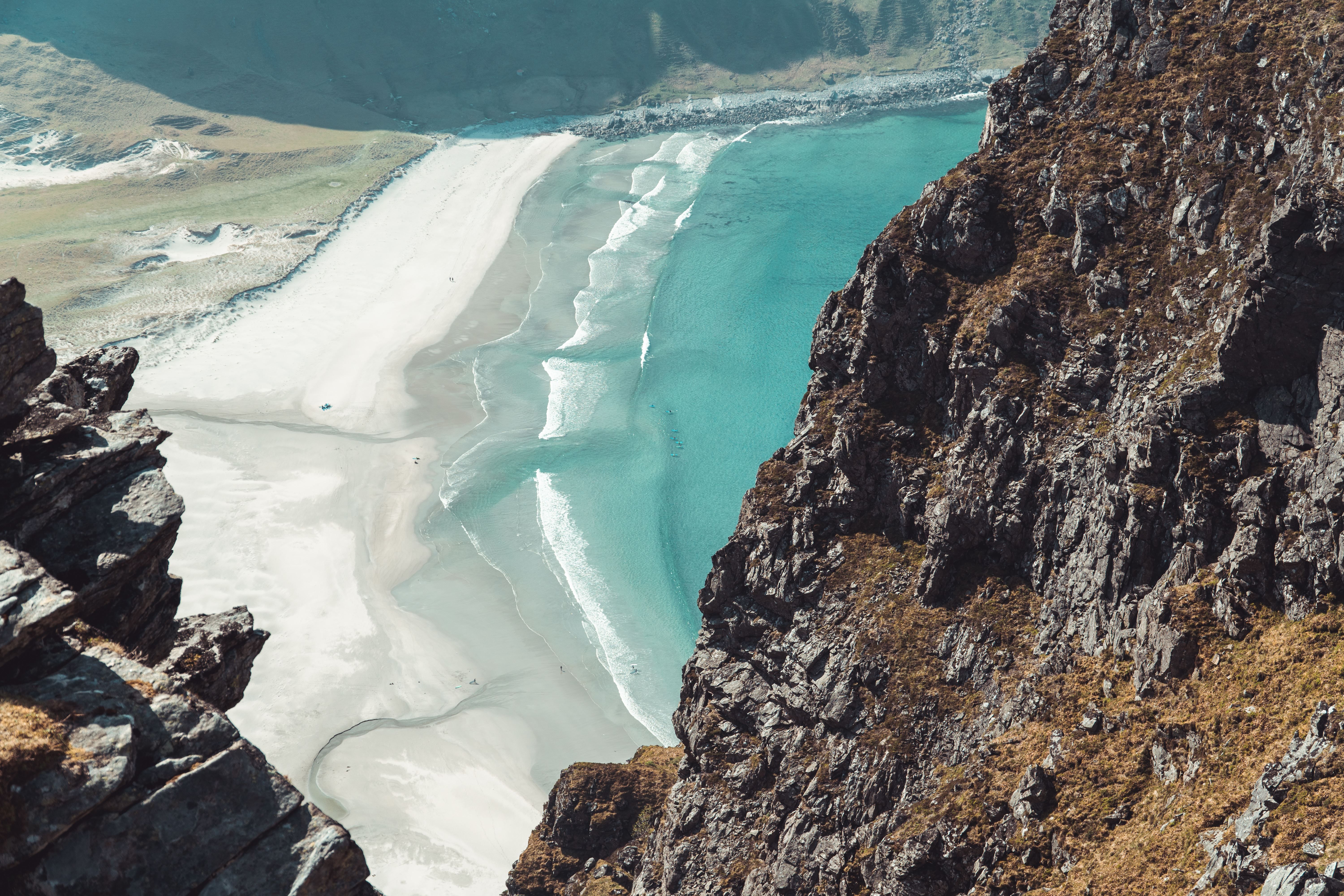 High-angle view from rocky cliffs over a crescent white-sand beach with turquoise water and breaking waves..
