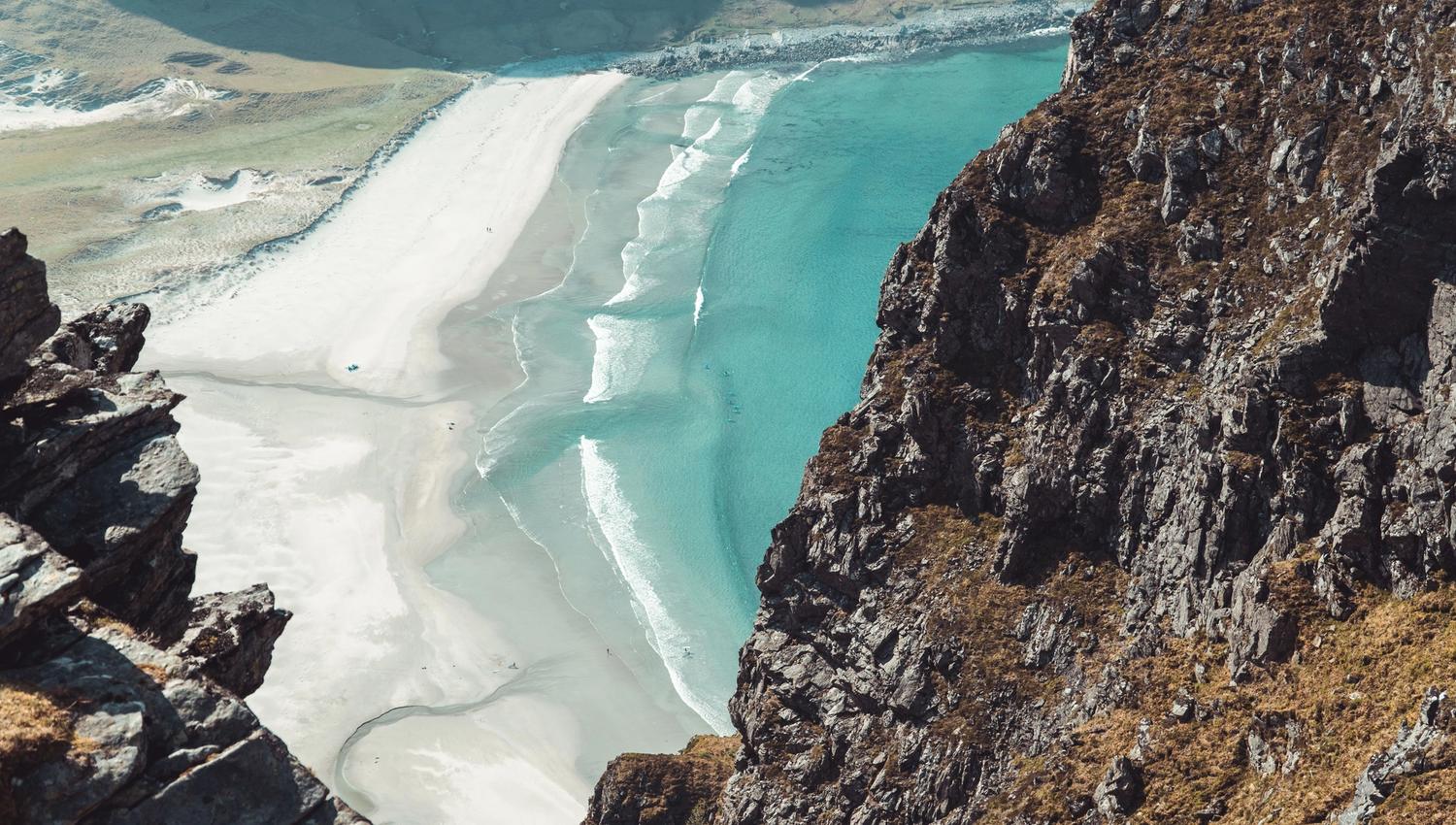 High-angle view from rocky cliffs over a crescent white-sand beach with turquoise water and breaking waves..