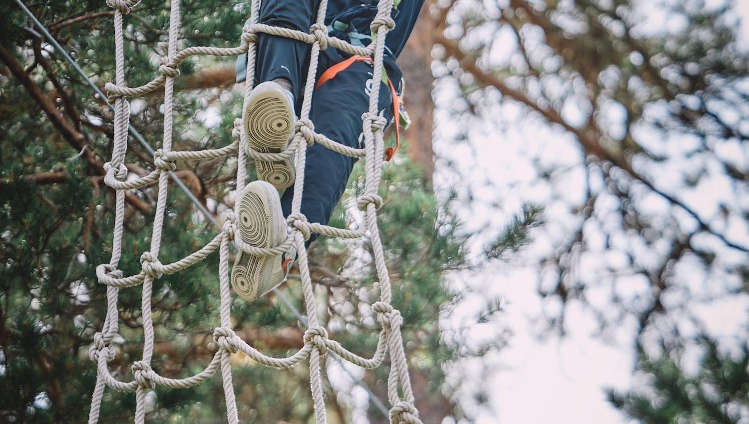 Ein person klatrar i eit tau-nett i ein skogkledd klatrepark, sikra med sele og line mellom trea..