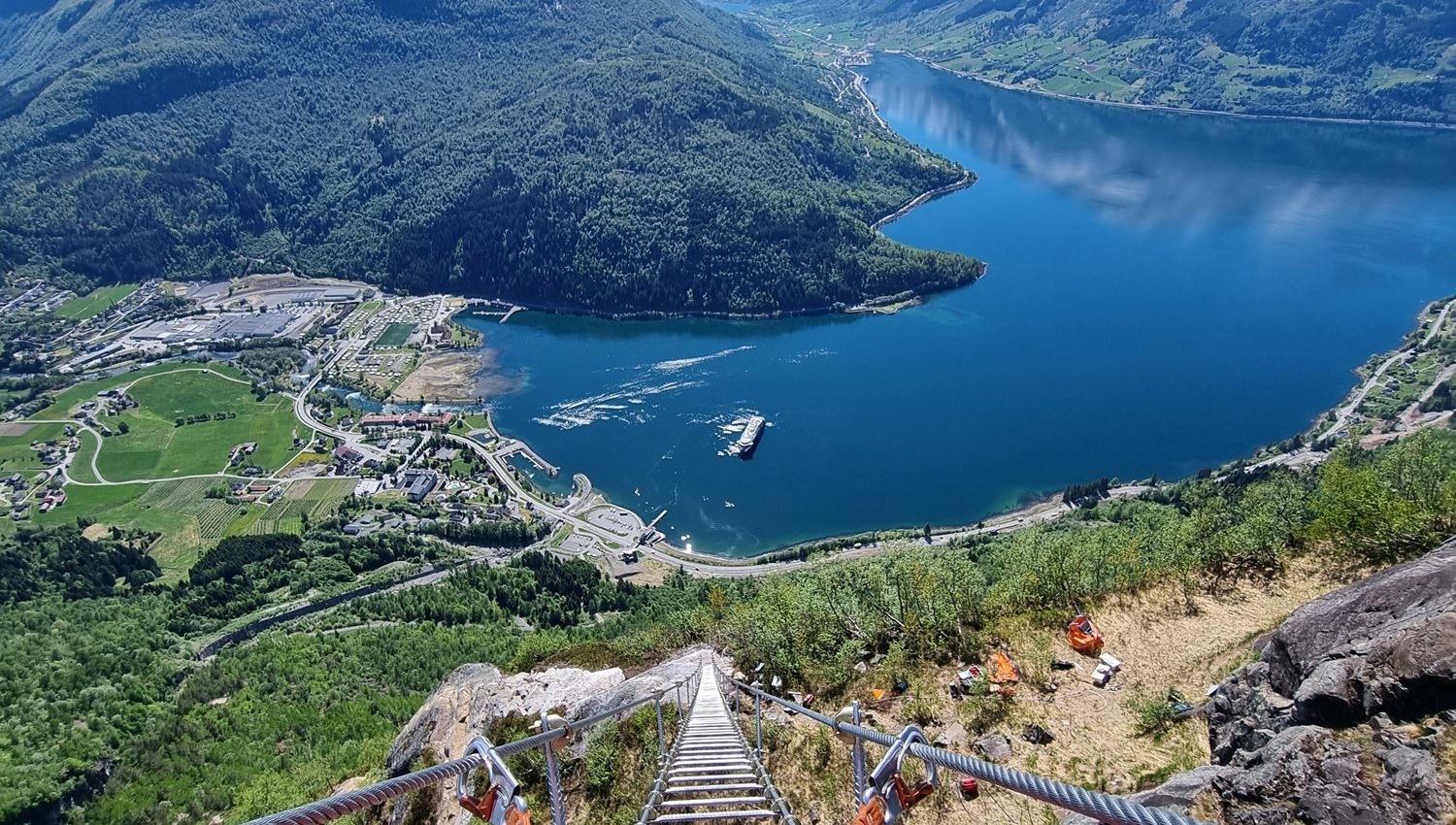 Utsikt ned ei stige med vaierar frå fjellside mot ein djup blå fjord, grøne dalar og snødekte fjell..