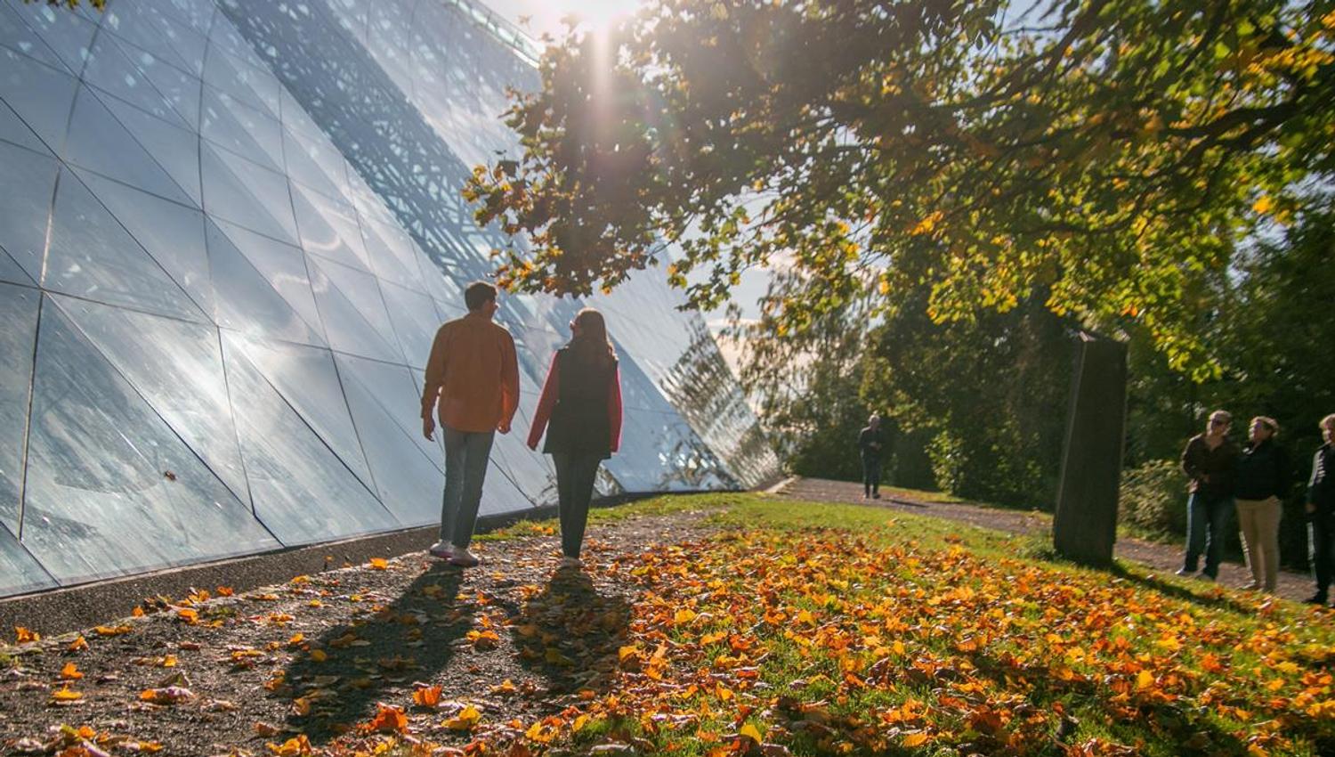 To personer går på en gressbakke med høstløv, ved siden av en stor bygning med glassfasade.
