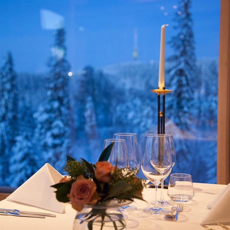 Elegant dekket bord ved vinduet med levende lys og vinterlig skoglandskap utenfor.