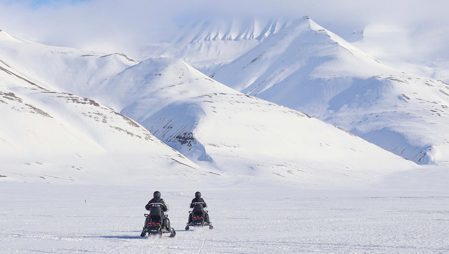 To personer på elektriske snøscootere i forgrunnen som kjører over en frossen tilsnødd slette mot snødekte fjell i bakgrunnen