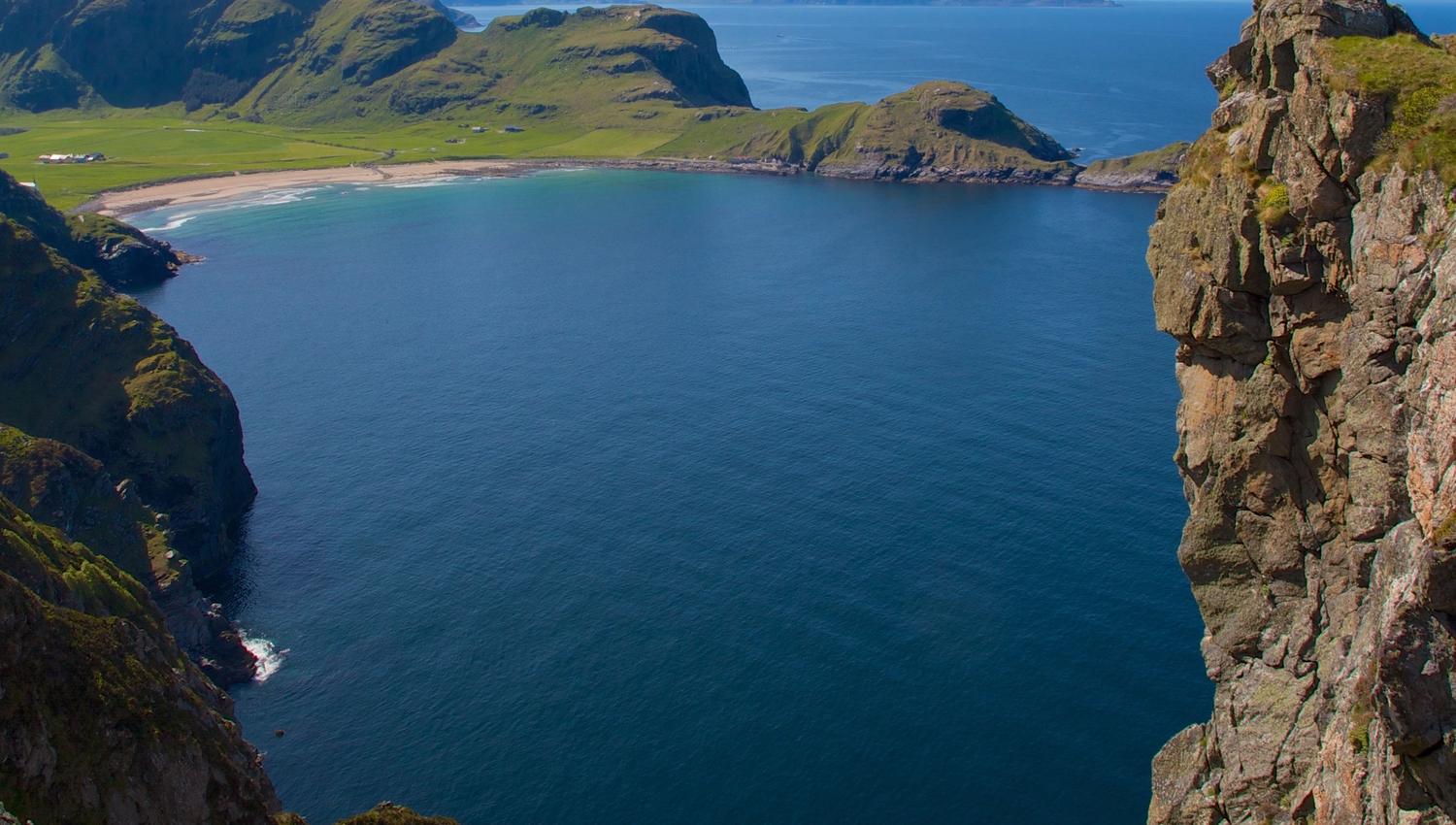Ein person i grøn jakke står på ei høg klippe i Ervik og ser utover ei blå havbukt med fjell og strand..