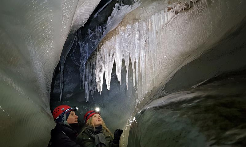to personer med hodelykt og hjelm som beundrer formasjonene i taket på isgrotten.
