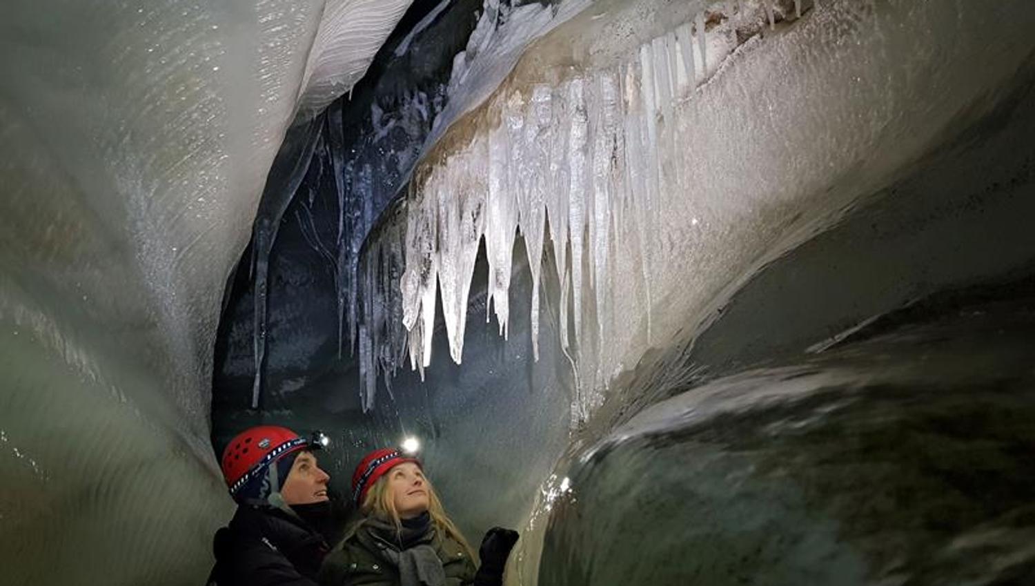 to personer med hodelykt og hjelm som beundrer formasjonene i taket på isgrotten.